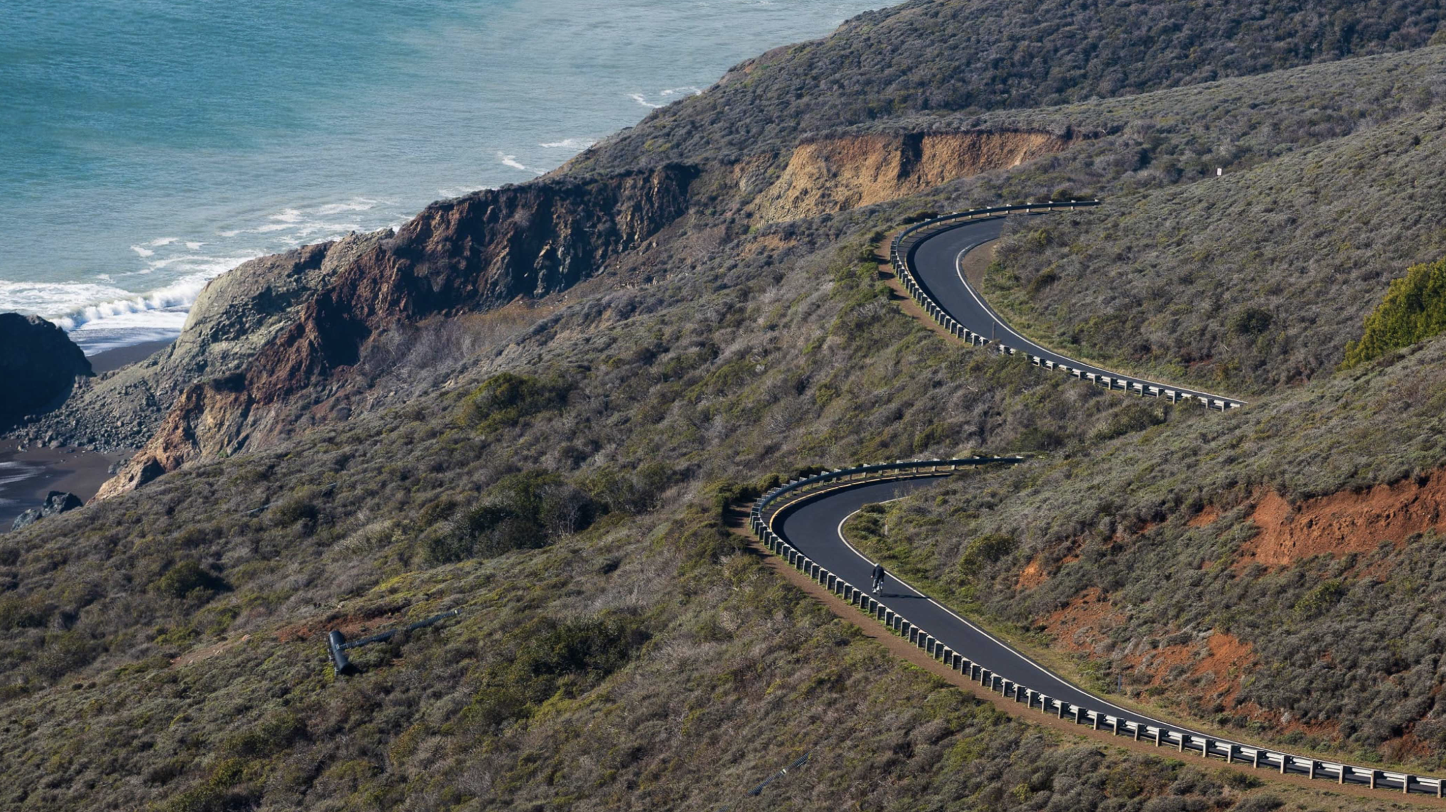 Winding coastal road on a hillside overlooking the ocean with waves crashing against rocky cliffs.