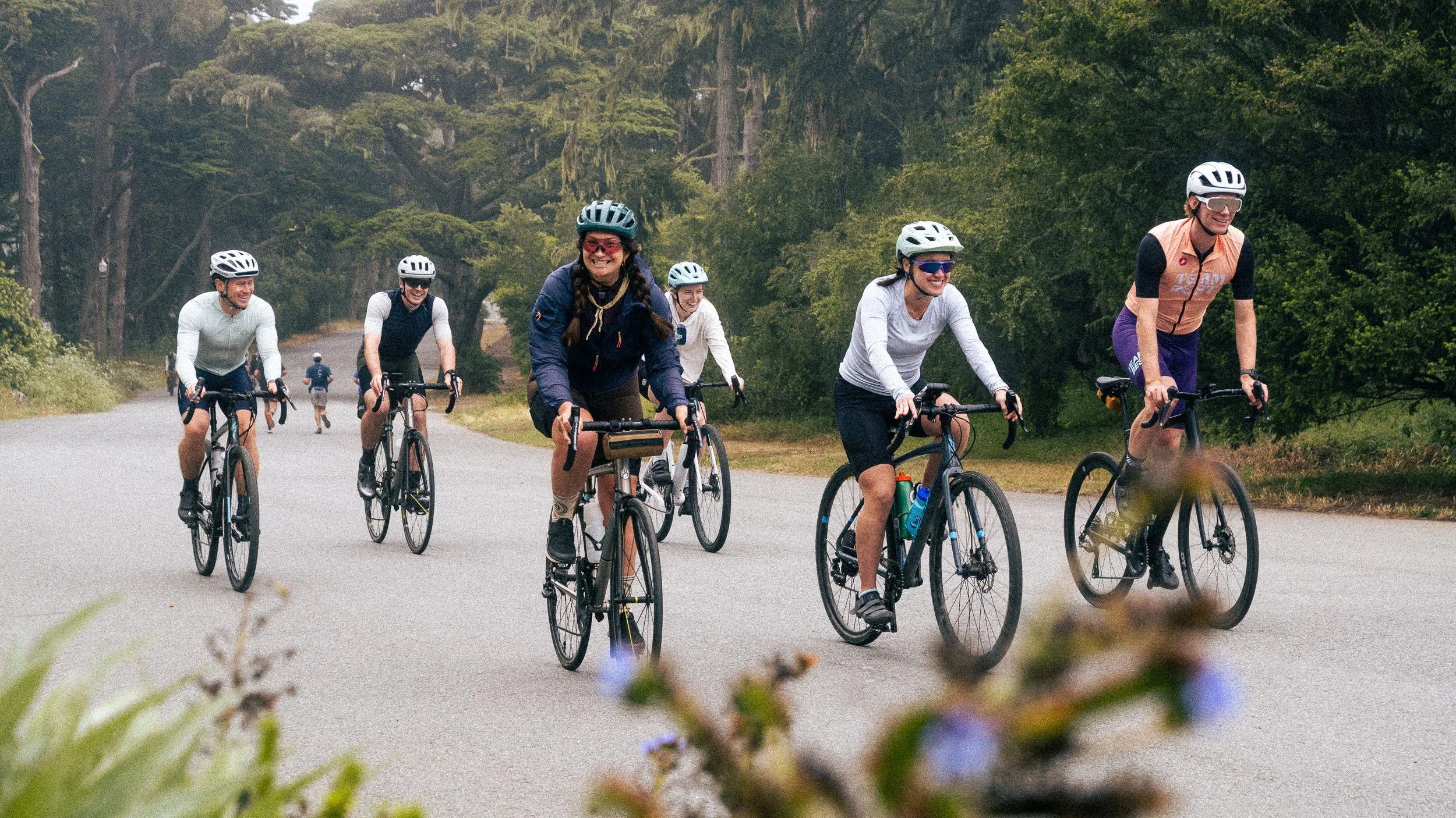 Group of six people riding bikes on a road through a wooded area, smiling and enjoying cycling day.