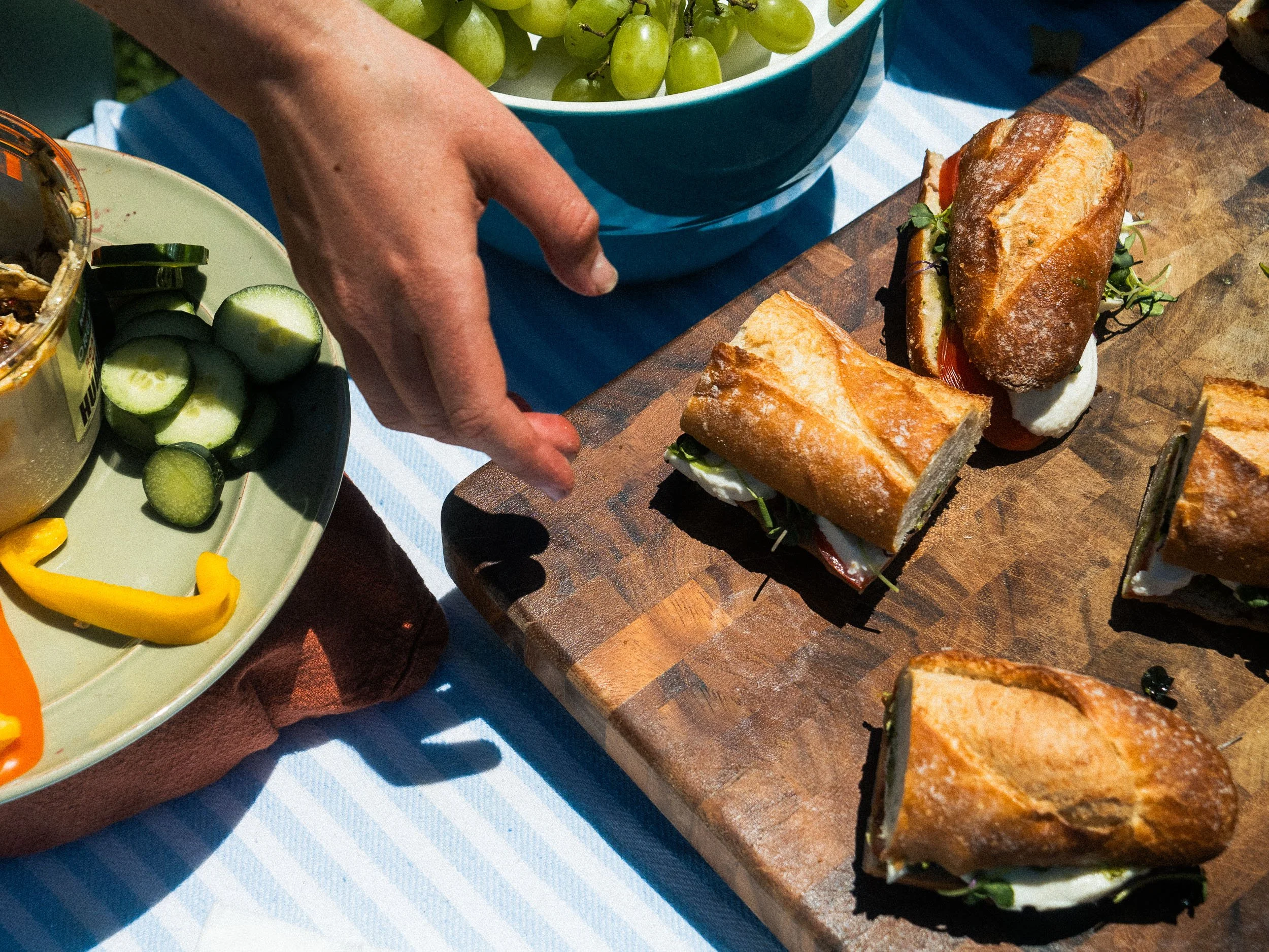 Close-up of a person's hand reaching for a small sandwich on a wooden cutting board, with bowls of green grapes, sliced cucumbers, and cheese nearby on a table with a blue and white striped tablecloth.