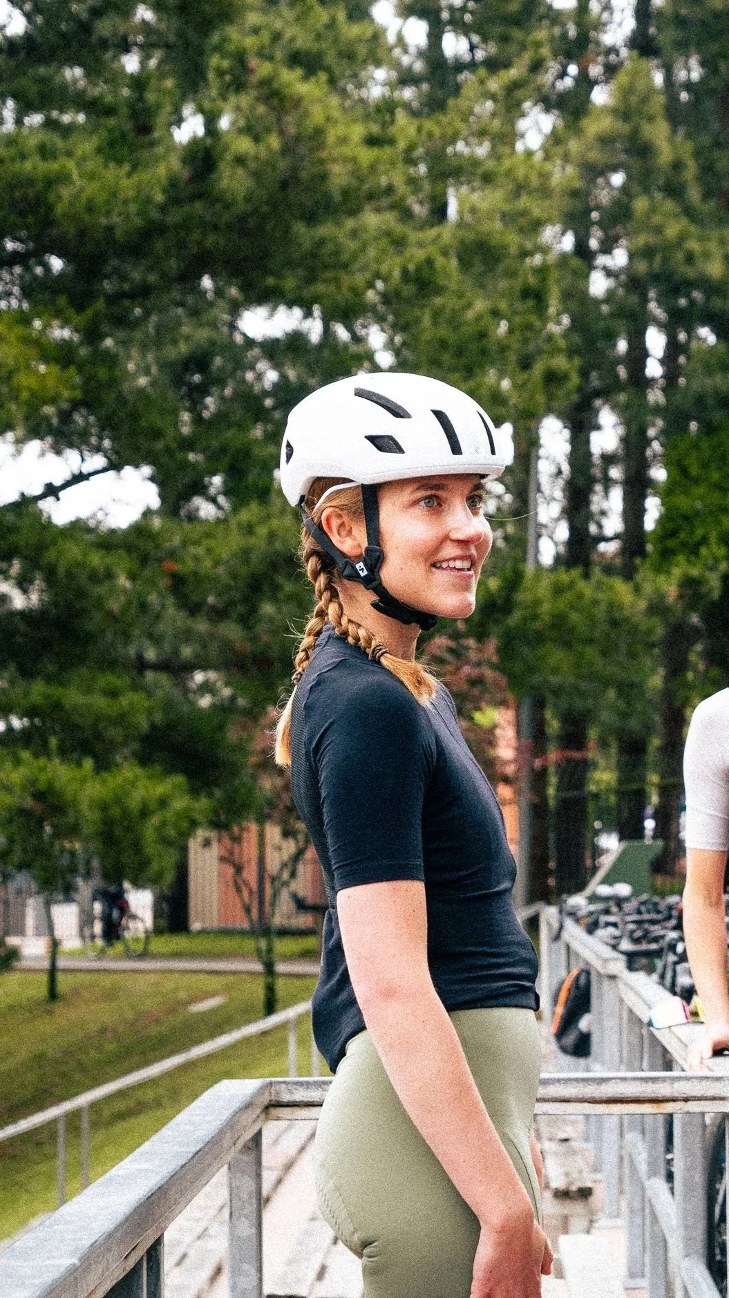 Woman wearing a white cycling helmet, black athletic shirt, and beige pants, standing outdoors on a wooden deck with trees and bicycles in the background.