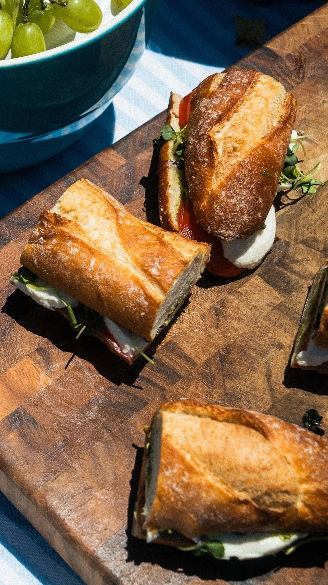 A close-up of a wooden serving board holding sliced sandwiches with crusty bread, lettuce, tomato, cheese, and other ingredients. In the background, there is a bowl of green grapes.