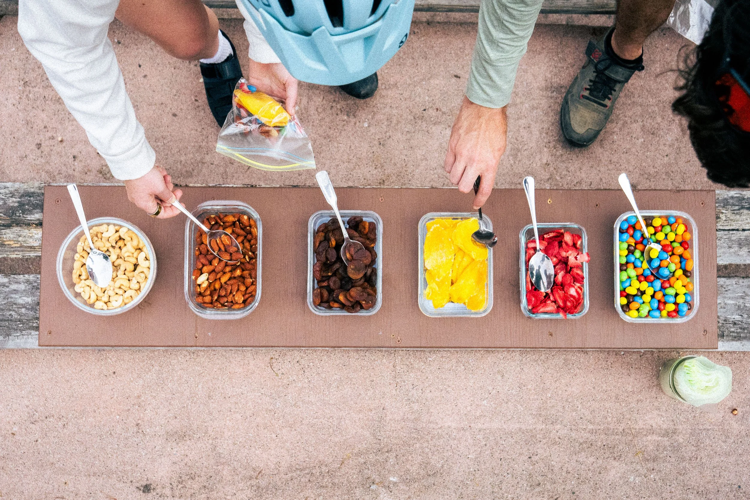 Top view of a table with various containers of colorful candies, including gummy worms, chocolate-covered candies, and fruit-flavored candies, with two people serving themselves.
