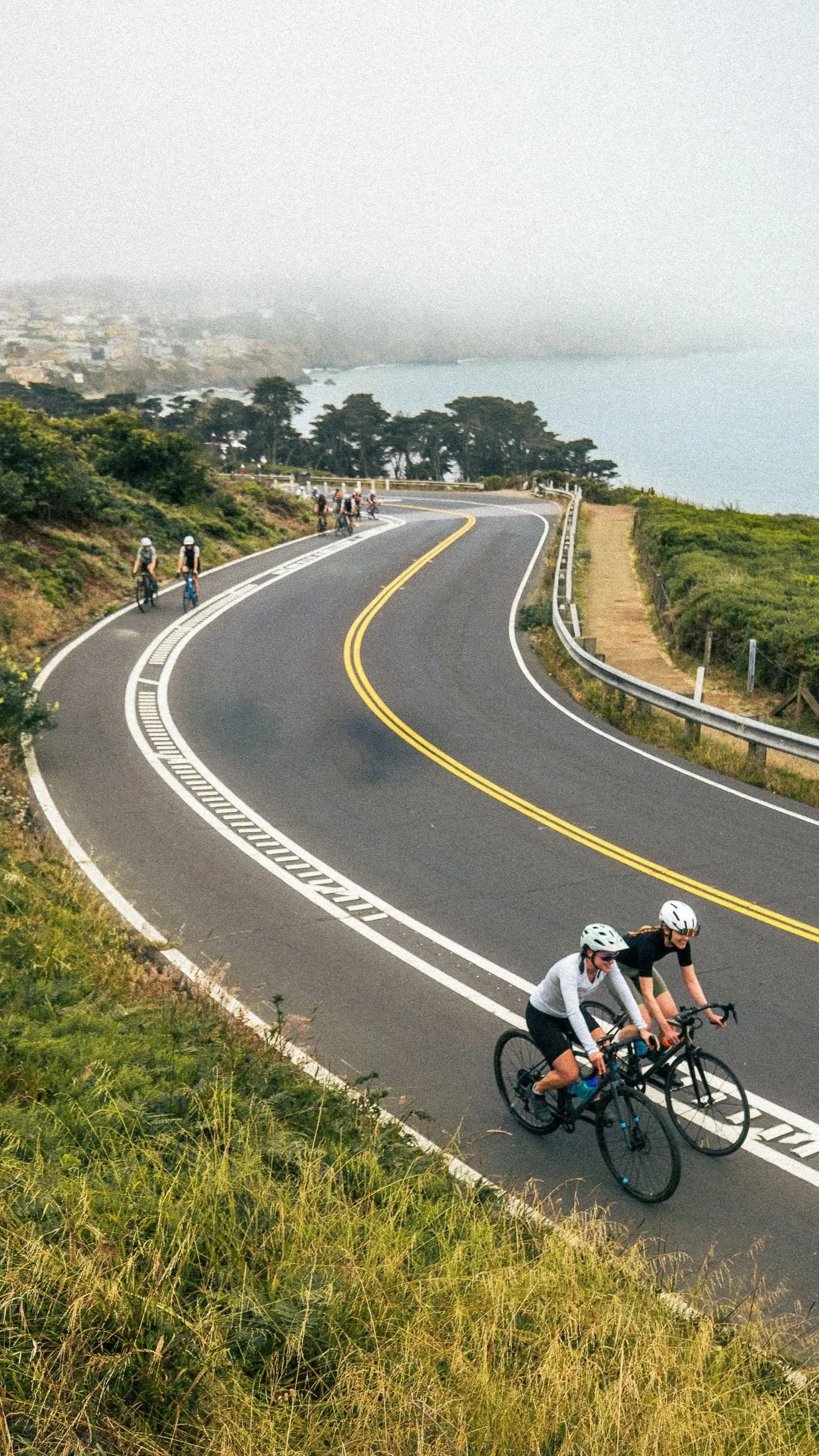 Cyclists riding on a winding coastal road with green vegetation and ocean view in the background.