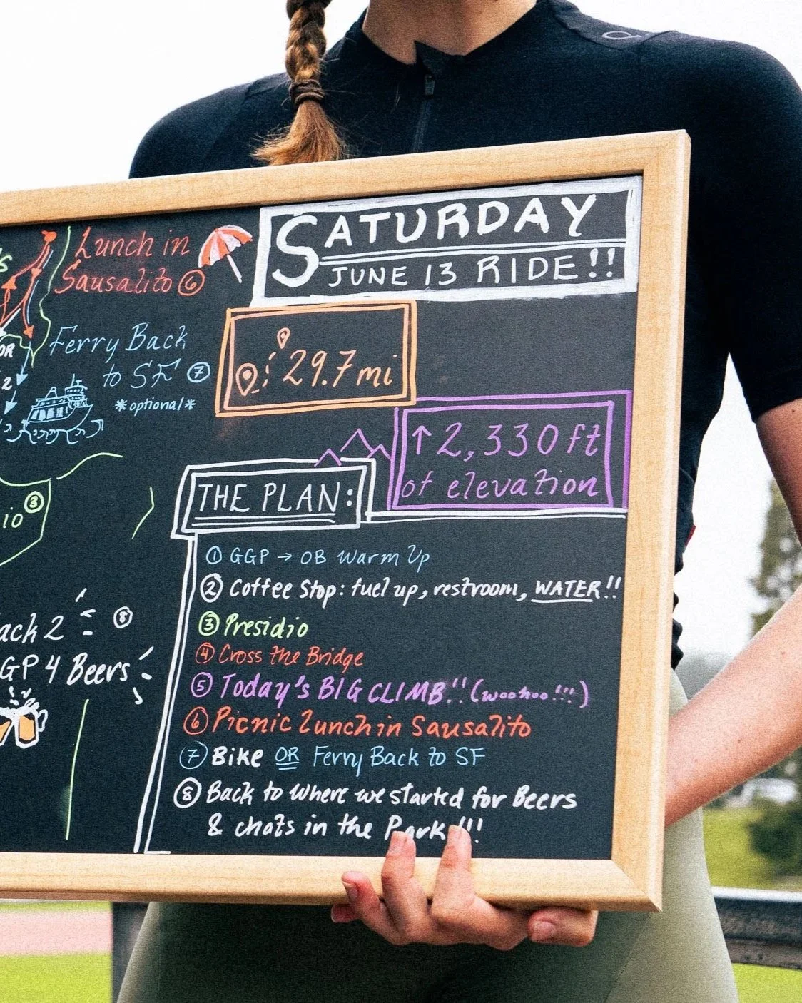 A person holding a blackboard with handwritten notes about a Saturday bike ride on June 13. The notes include a route map, distance of 29.7 miles, elevation gain of 2,330 feet, and a detailed plan with stops at a coffee shop, crossing a bridge, climbing, a picnic lunch, and returning to San Francisco.