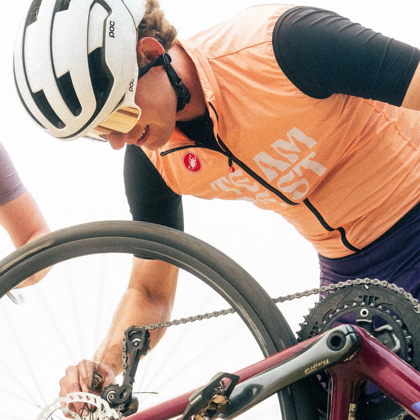 A cyclist wearing a white helmet and orange jacket adjusting their bike gear.