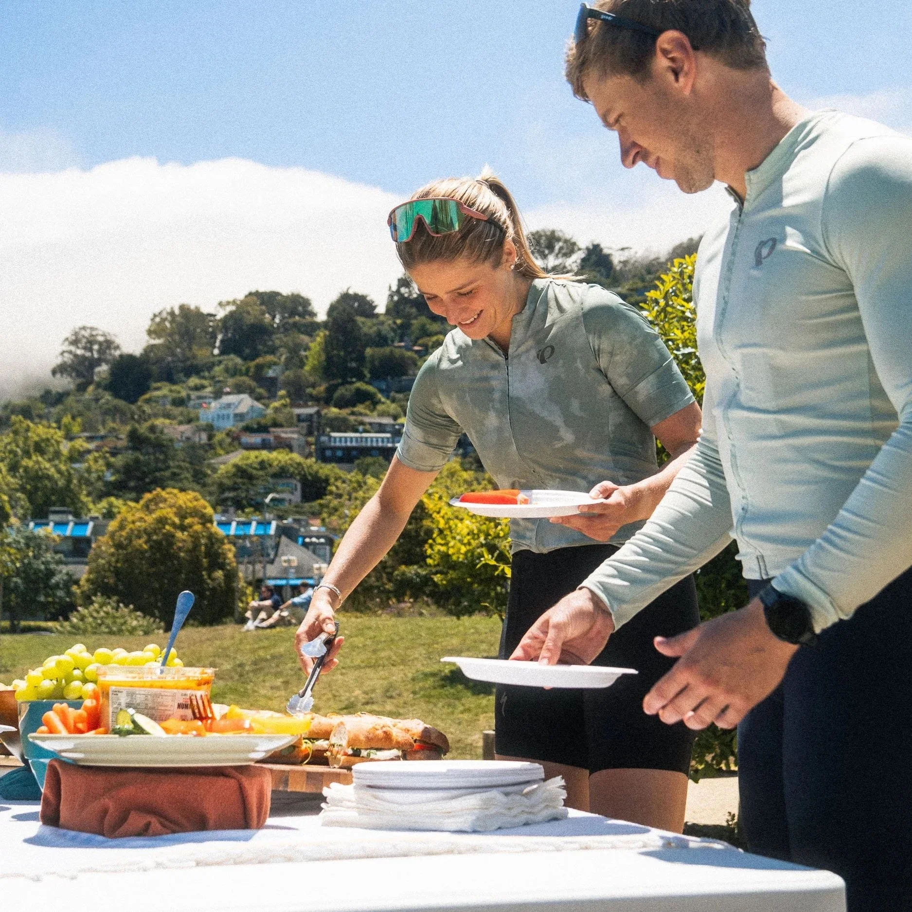 People serving themselves food at an outdoor buffet table with fruit, vegetables, and plates, during a sunny day with blue skies.