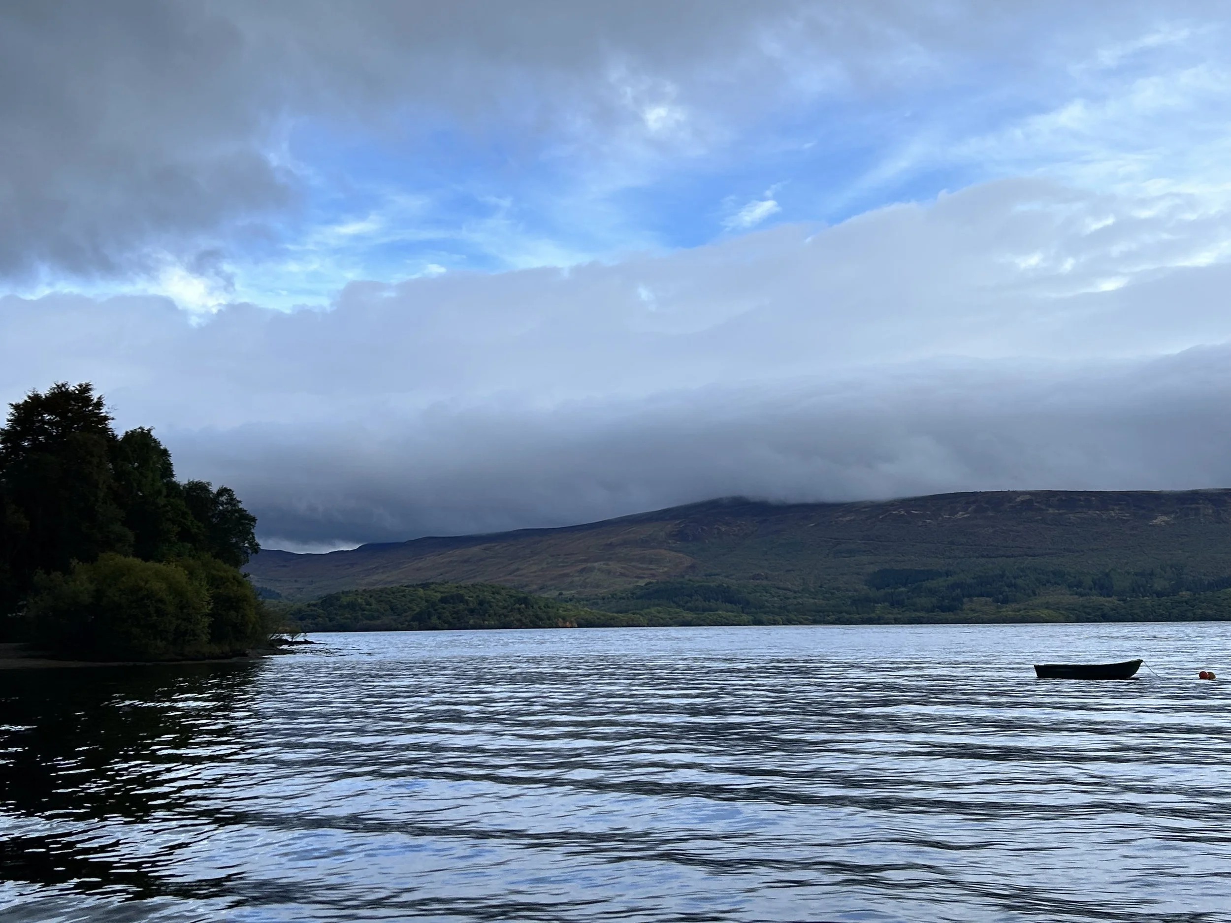 A lake with a small boat floating on it, surrounded by green trees on the left and rolling hills in the distance, under a mostly cloudy sky with patches of blue.