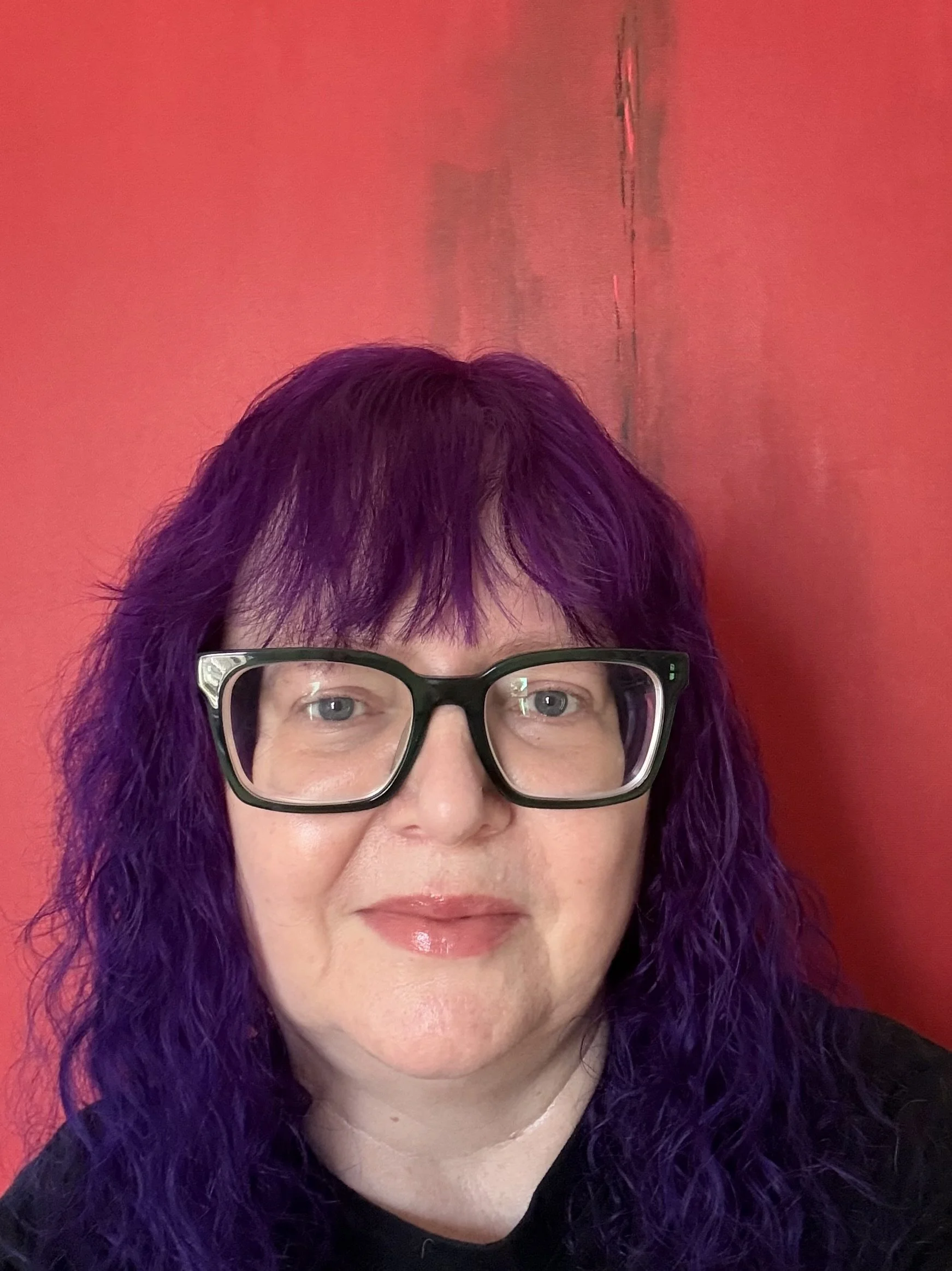 A close-up of a woman's face with purple hair, wearing glasses, against a red background.
