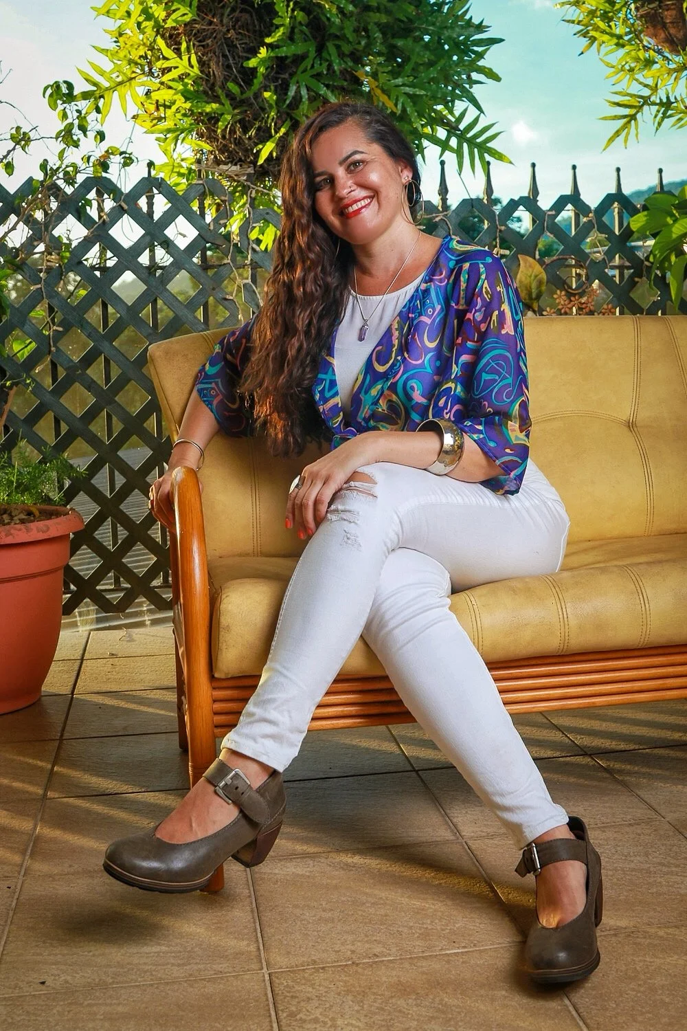 A woman with long wavy hair sitting on a yellow outdoor sofa, smiling at the camera. She is wearing a colorful patterned blouse, white ripped jeans, and gray buckle shoes. Behind her are greenery, a lattice fence, and a partly cloudy sky.