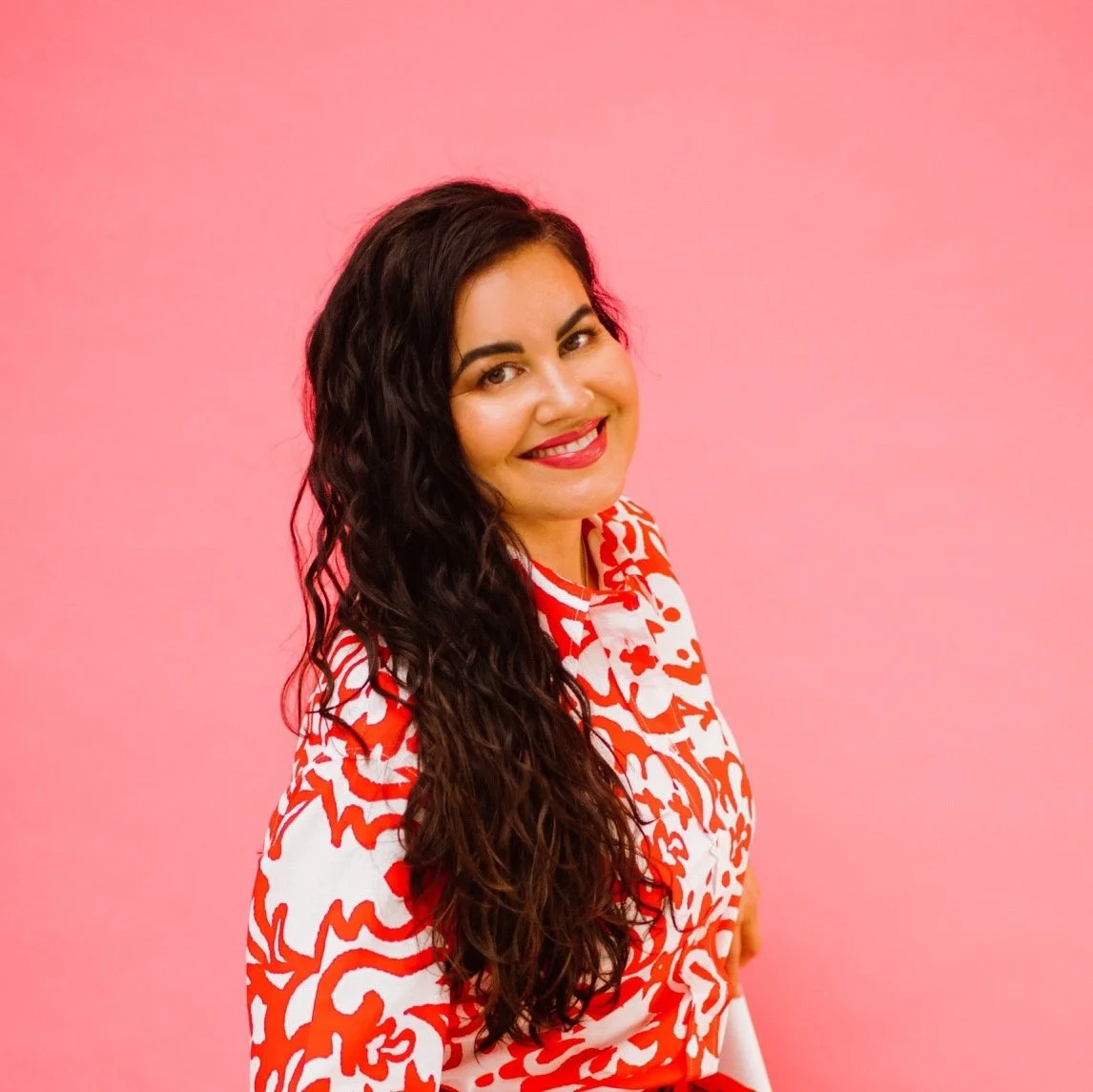 A woman with long, curly dark hair, wearing a red and white patterned top, smiling against a pink background.