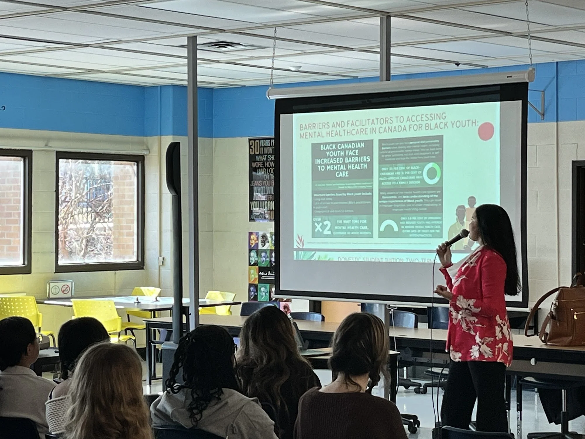 A woman in a red floral dress speaking into a microphone during a presentation in a classroom. A slide with text about barriers and facilitators to accessing mental healthcare in Canada for Black youth is projected behind her. Students are seated and listening, with some windows letting in natural light.