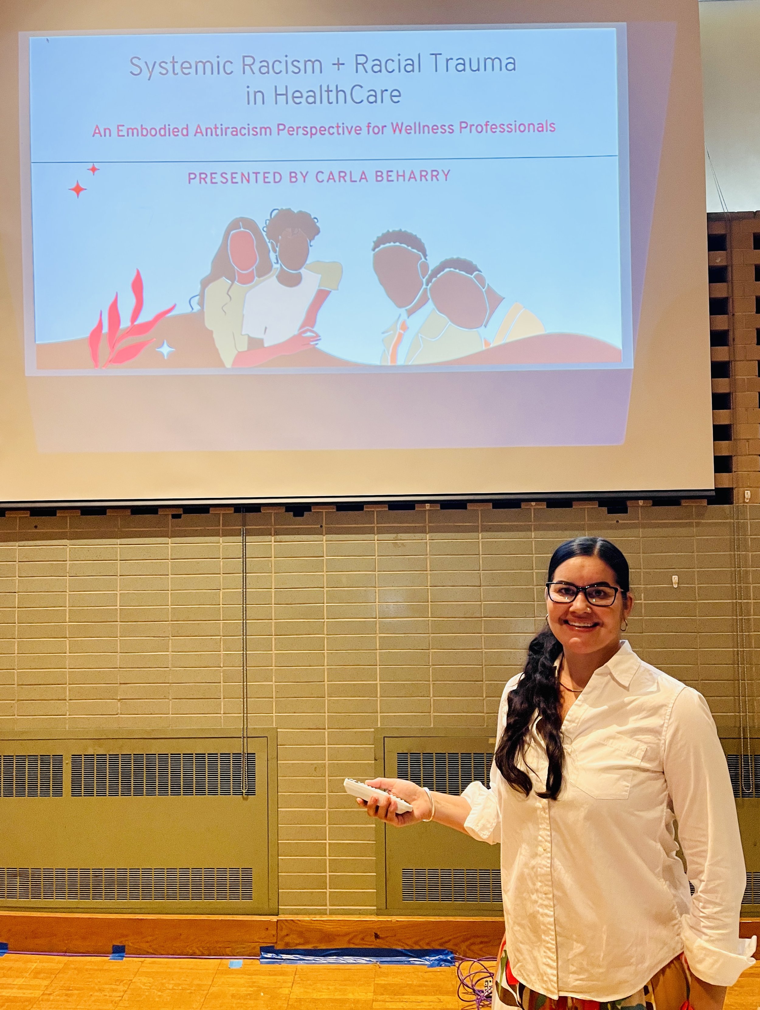 A woman in a white shirt holding a phone, standing in front of a presentation slide titled 'Systemic Racism + Racial Trauma in Health Care' and 'An Embodied Antiracism Perspective for Wellness Professionals,' with illustrations of four diverse people on the slide.