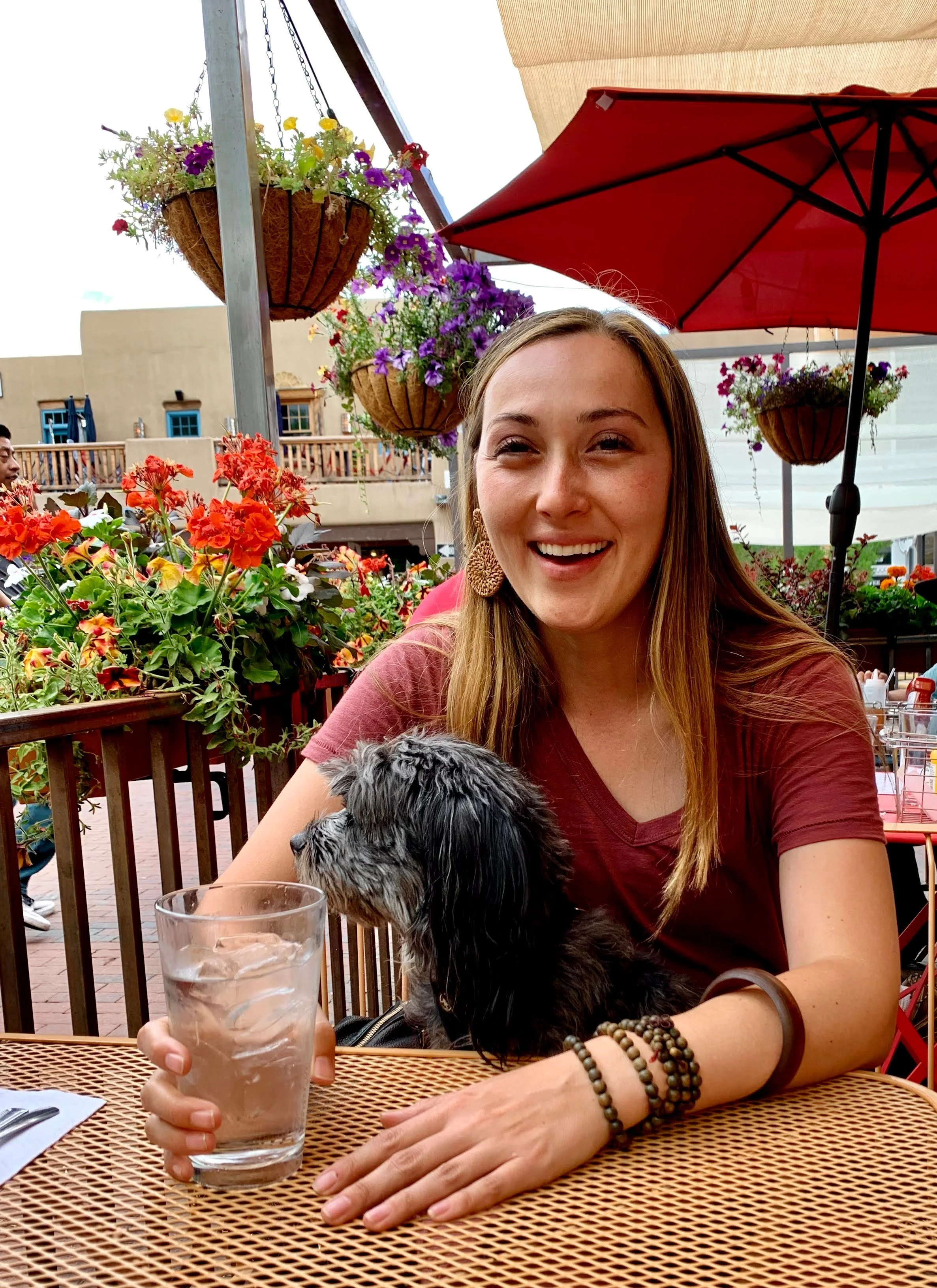 A woman with long blonde hair wearing a burgundy shirt and multiple bracelets on her right wrist, holding a glass of water with ice. She is sitting at a table outside under a red patio umbrella, with colorful flowers and hanging baskets in the background.