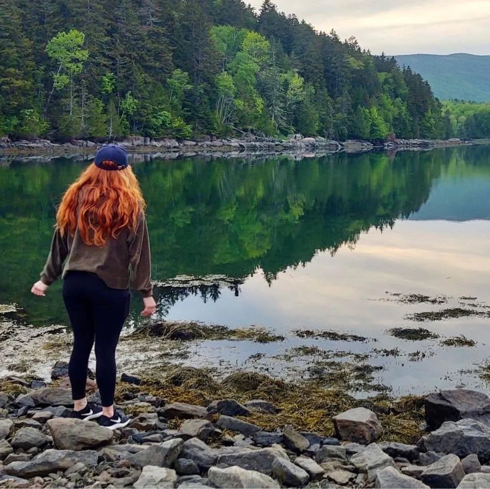 A woman with long red hair, wearing a baseball cap, dark leggings, and a jacket, stands on a rocky shoreline looking at a calm lake surrounded by green trees and distant mountains.