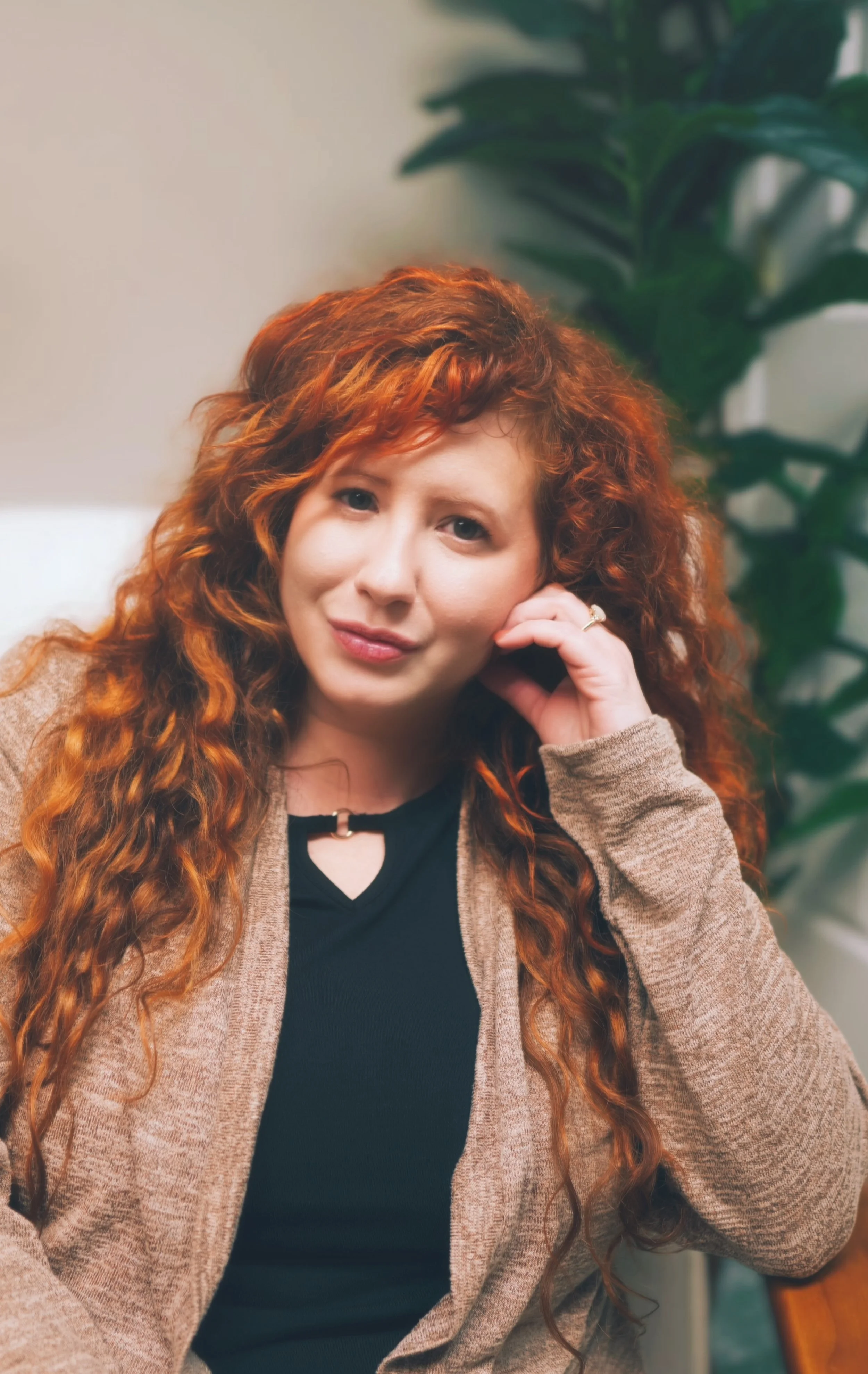 A woman with long, curly red hair and fair skin is sitting indoors, resting her chin on her hand, looking directly at the camera with a slight smile. She is wearing a black top and a light brown cardigan, and there is a large green leafy plant in the background.