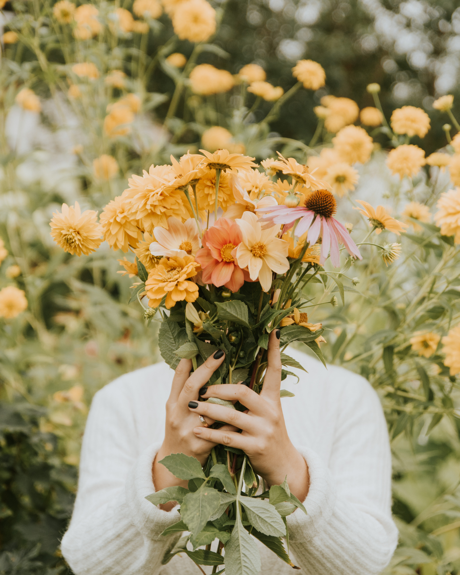 Person holding a bouquet of yellow and pink flowers in front of their face, with a blurred background of similar flowers.