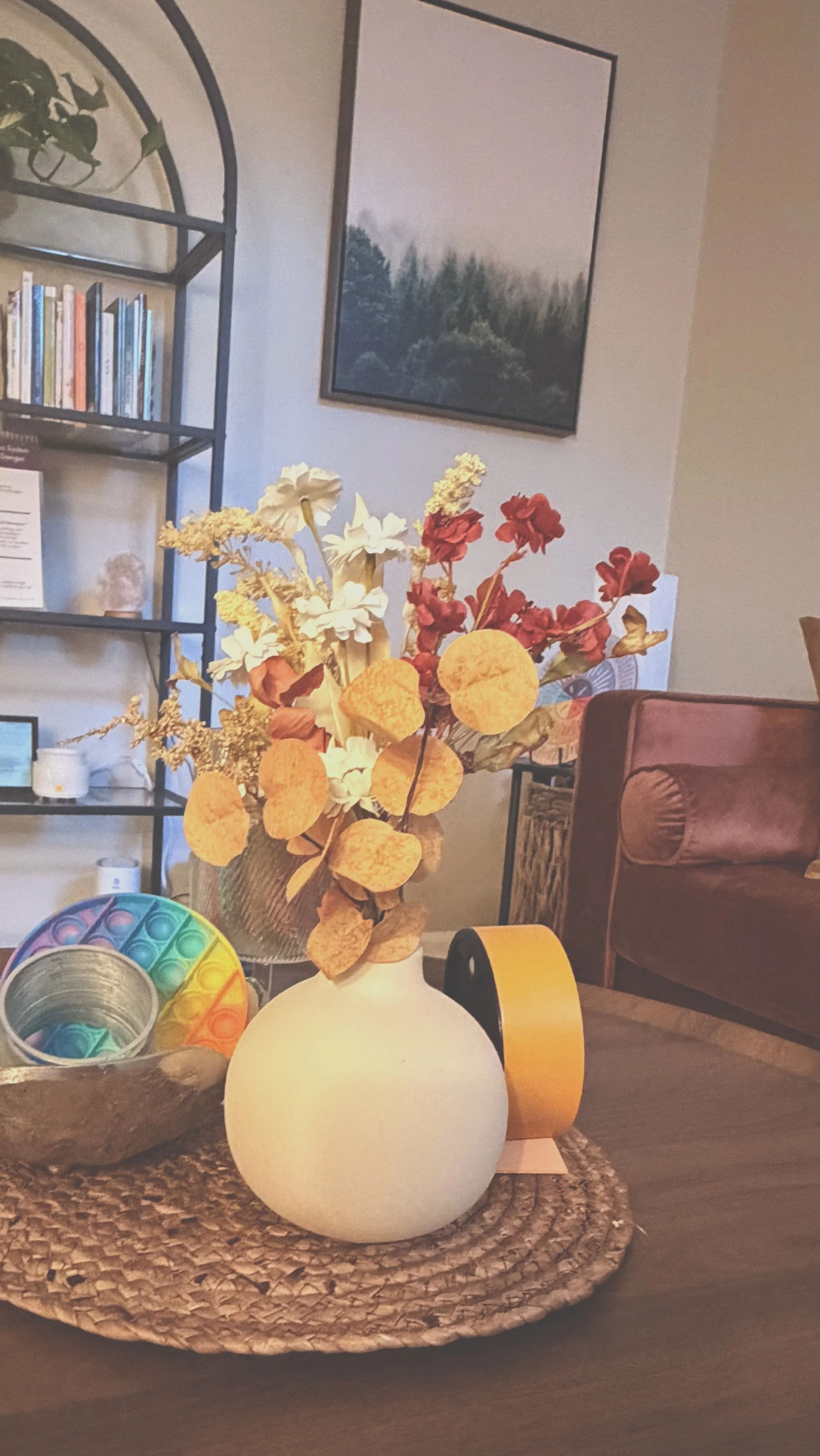 A beige spherical table lamp, a colorful rainbow pop-it toy, and a small metal tin on a woven placemat on a wooden table. In the background, a flower arrangement with yellow, white, and red flowers in a white vase, a bookshelf with books, a framed picture of a forest, and parts of a leather armchair are visible.