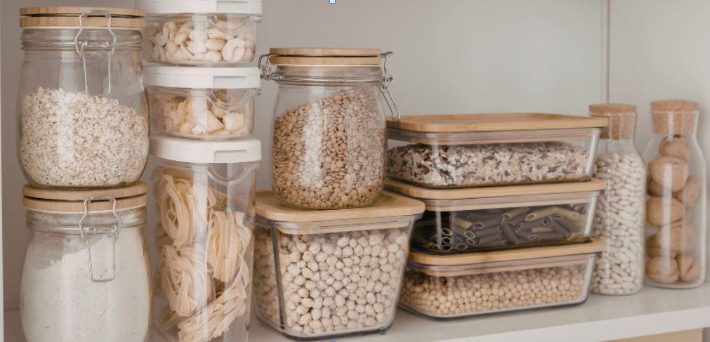 A variety of glass jars and containers filled with dried pasta, grains, and legumes are stored on a kitchen shelf.