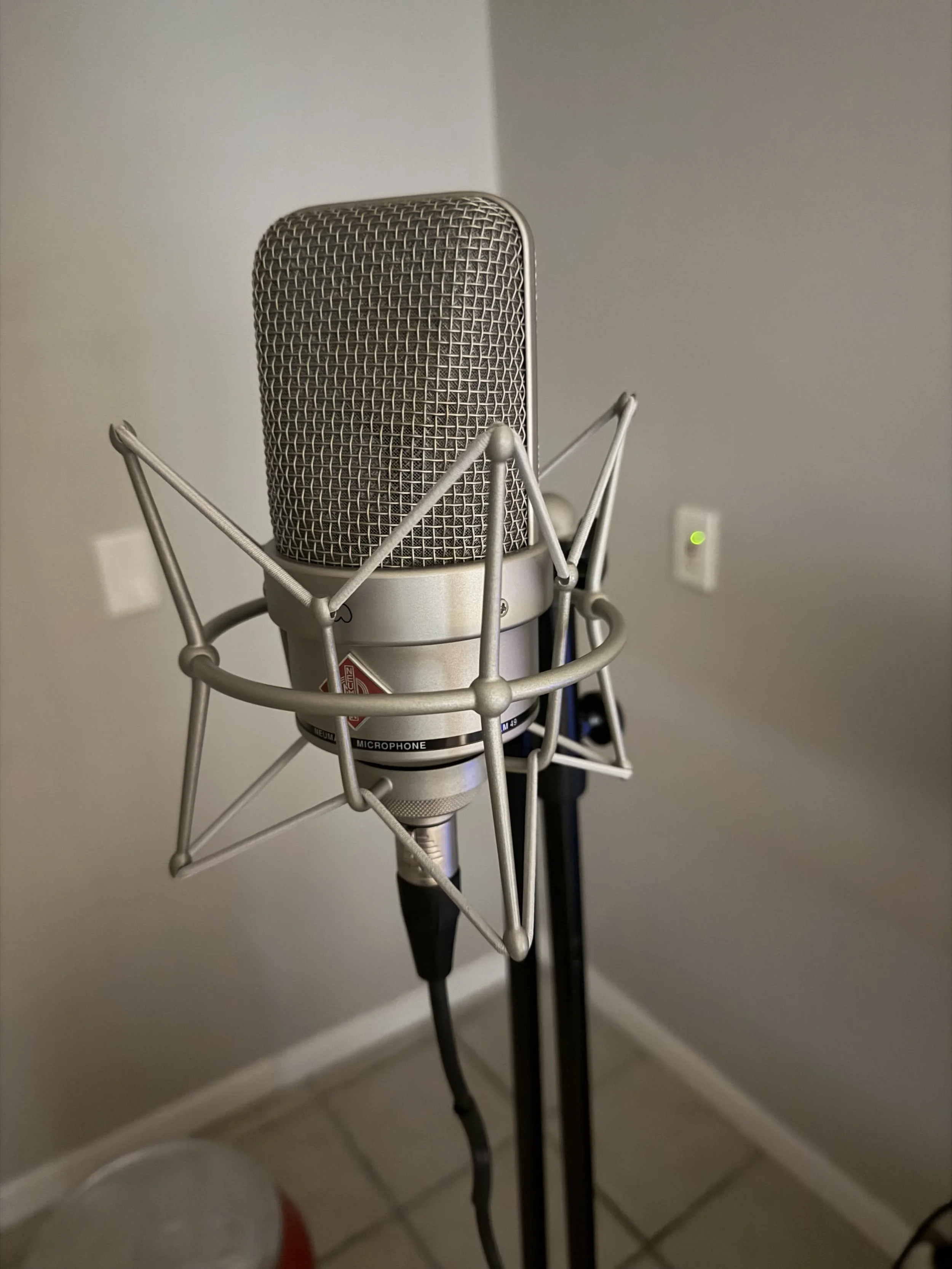 Close-up of a professional studio microphone suspended in a shock mount, with a gray wall and electrical outlets in the background.