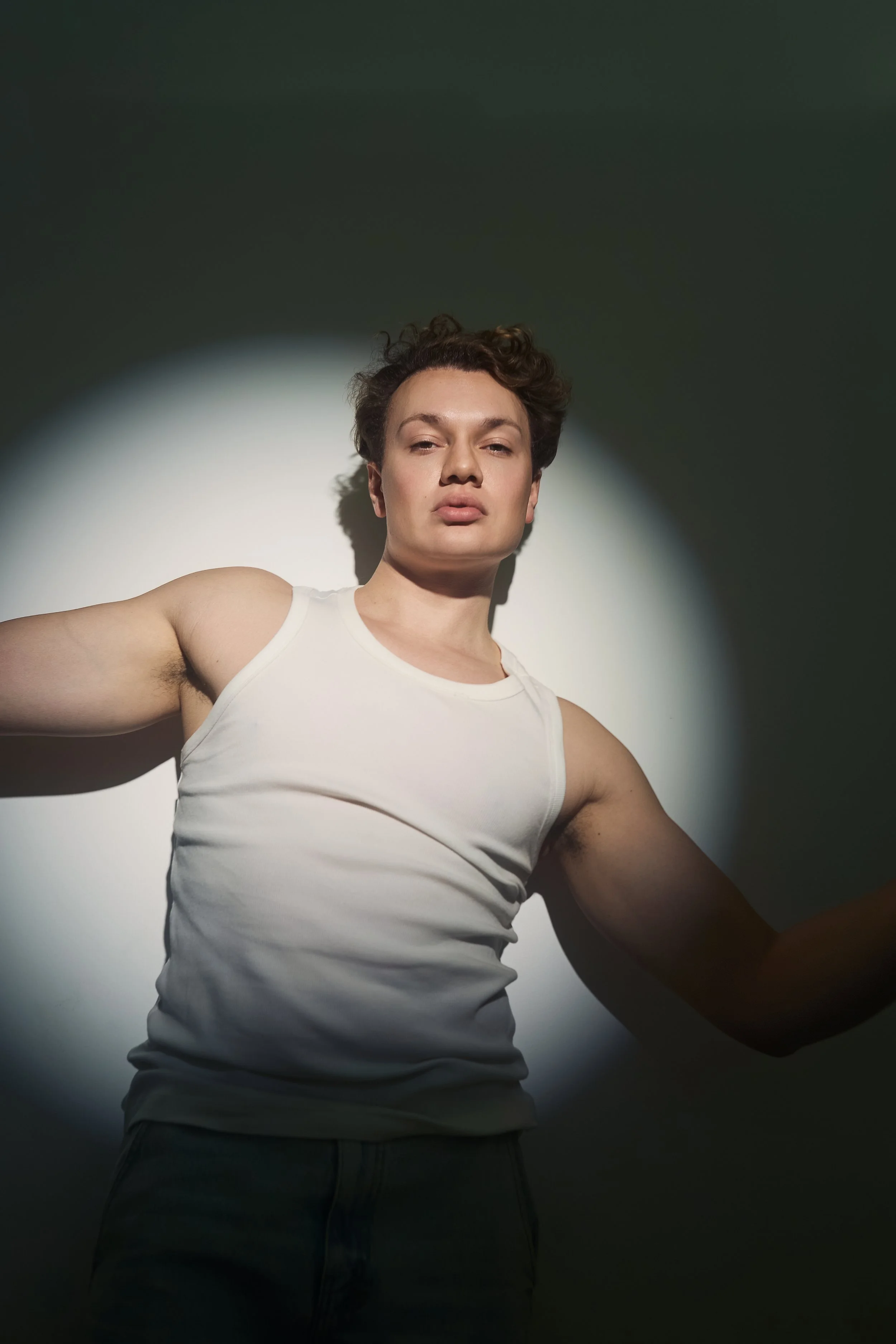 A woman taking a selfie against a dark background with a white light focused on her face and upper body. She has short, curly hair and is wearing a white tank top.