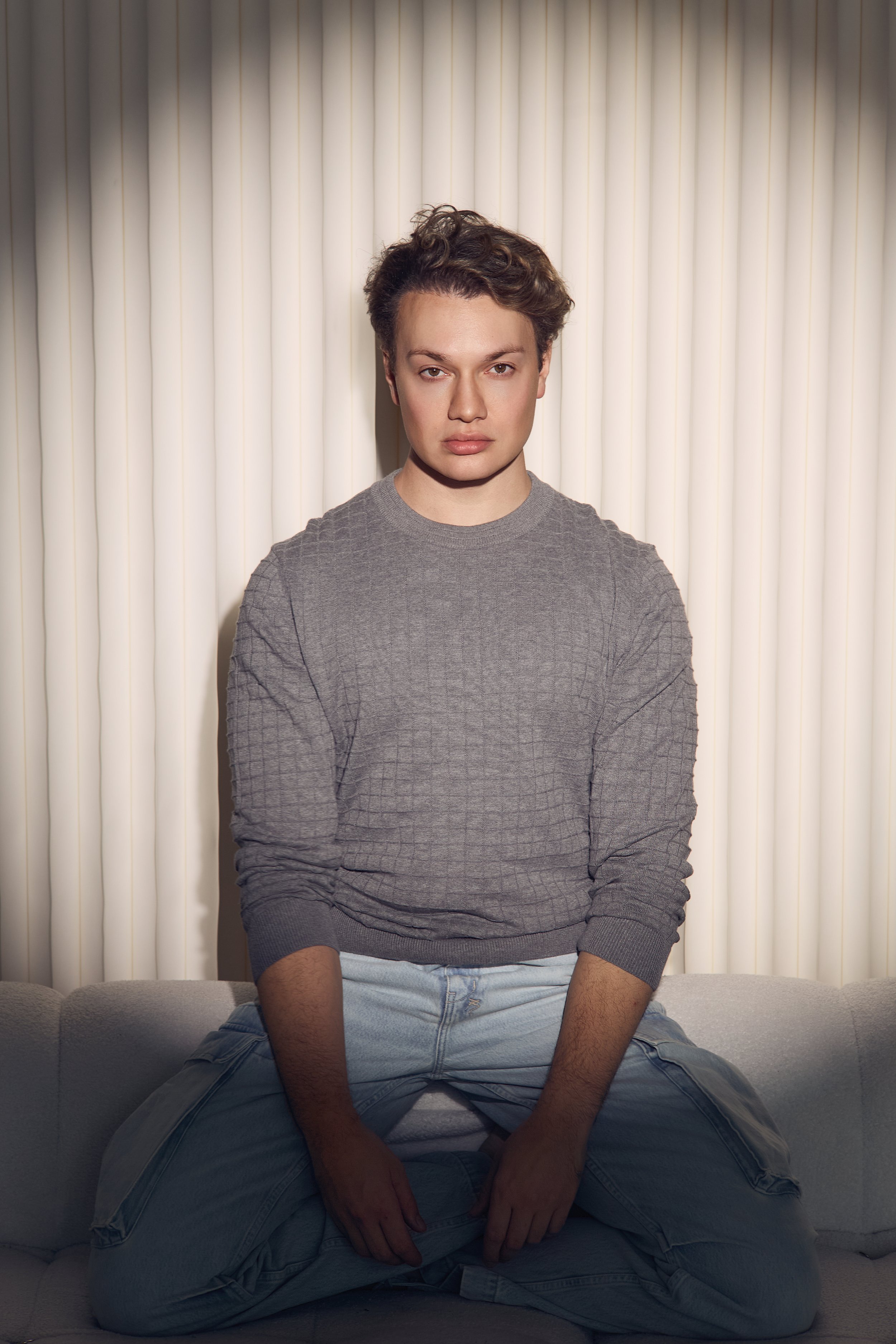 A young person with short, curly hair sitting cross-legged on a couch, wearing a gray long-sleeve shirt and light jeans, in front of a background with vertical white blinds.