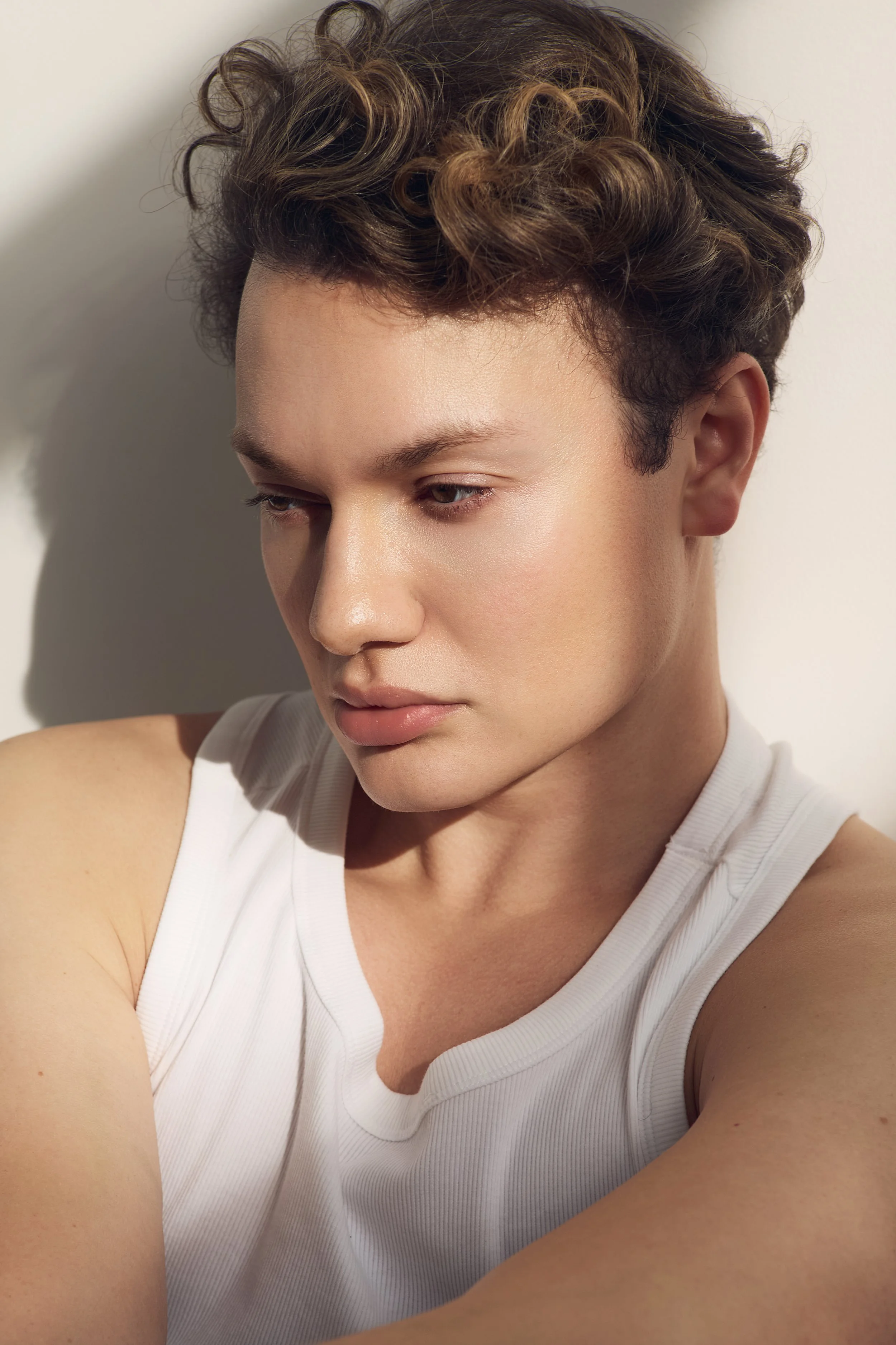 Close-up of a young woman with short, curly hair in shades of brown, wearing a white sleeveless top, looking down with a content expression.