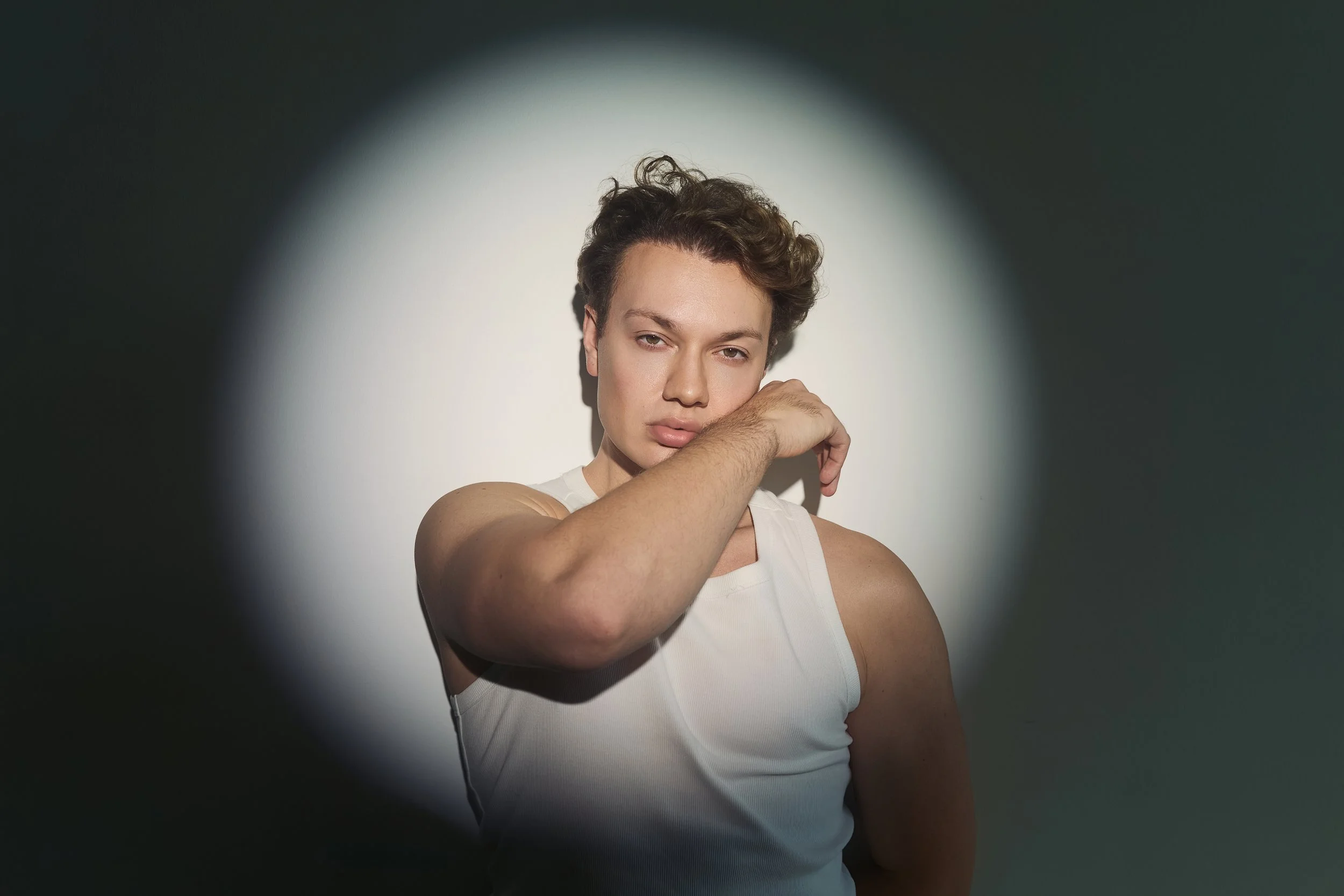 Portrait of a young person with short curly hair, wearing a white tank top, posing with one arm across their face, against a dark background with a focused light spot.