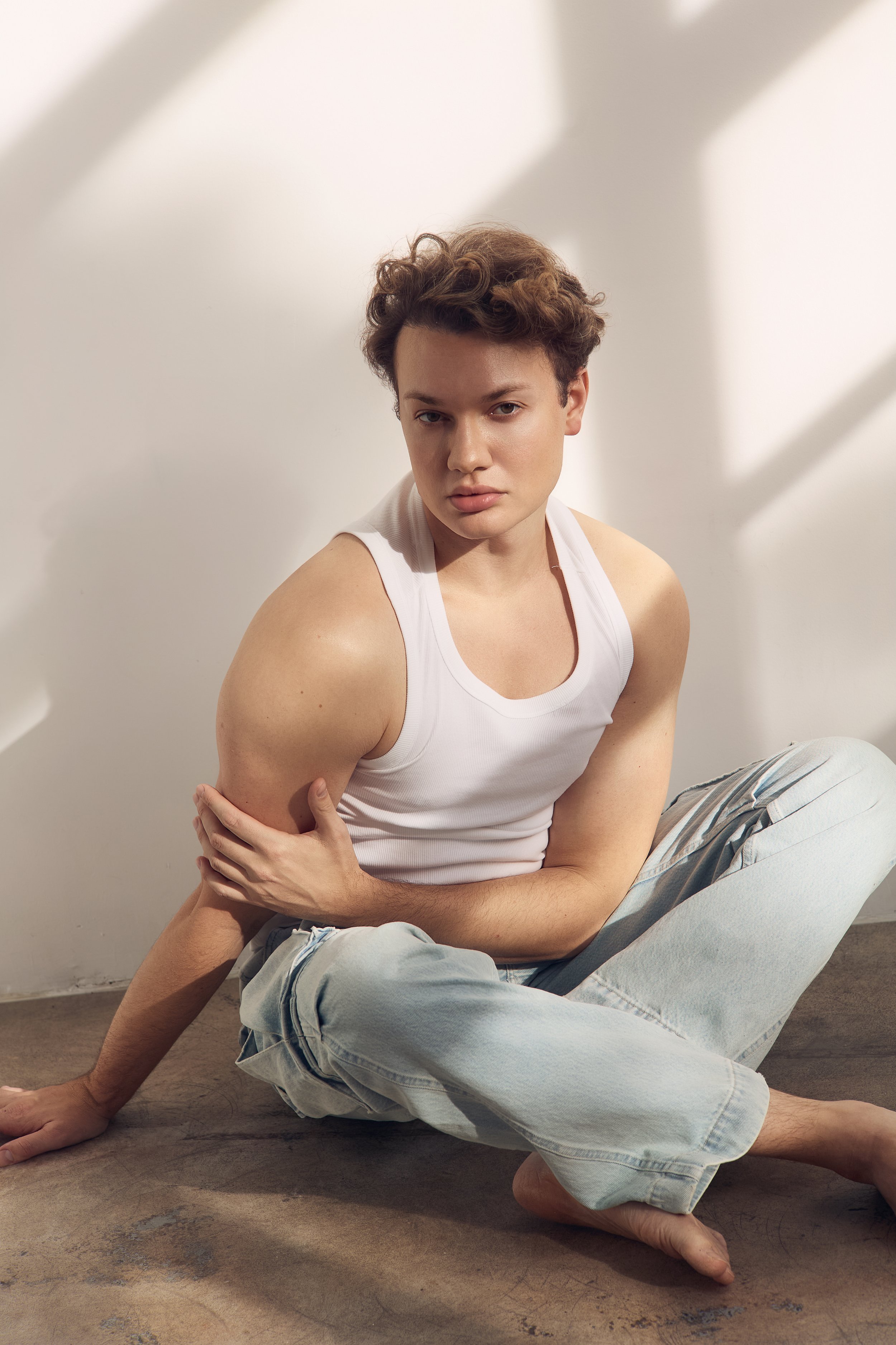 A person in a white tank top and light jeans is sitting on a wooden floor with one leg extended and the other bent, in a pose with their arm crossed over their chest, against a plain white wall background with soft shadows.