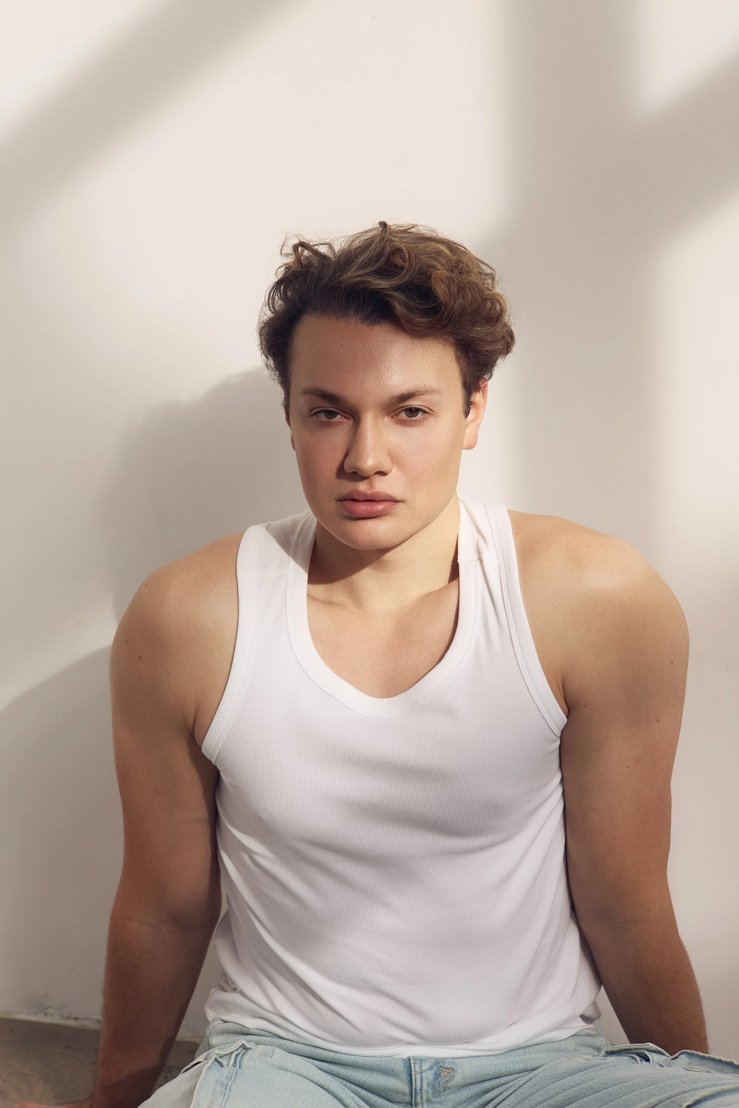 A young individual with short, curly brown hair and a fair complexion, wearing a white sleeveless top and light denim shorts, sitting against a white wall and looking at the camera.
