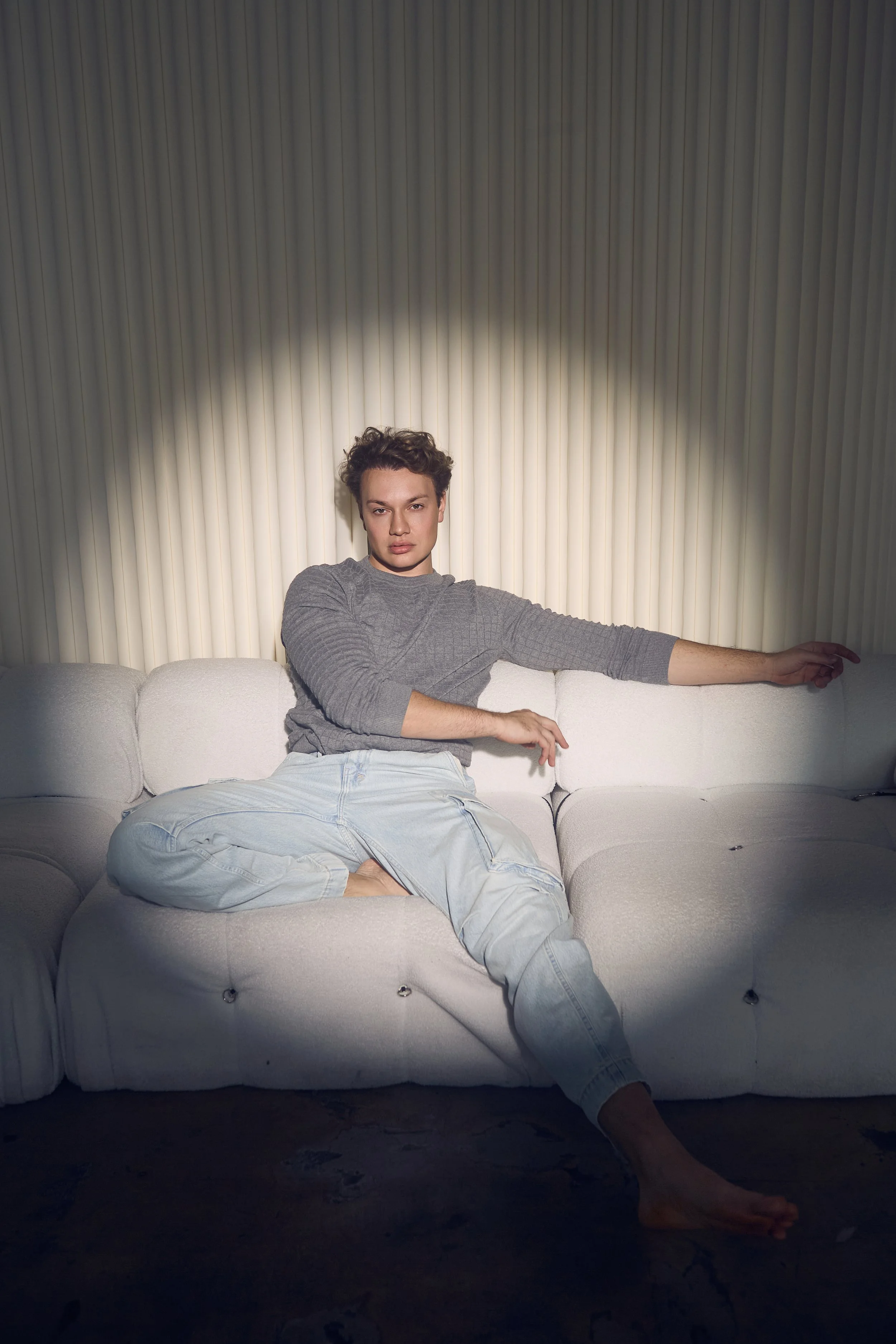 A young person with short, curly hair sitting on a white couch against a vertically striped wall, with one arm extended and the other resting on their lap, wearing a gray long-sleeve shirt and light-colored jeans.