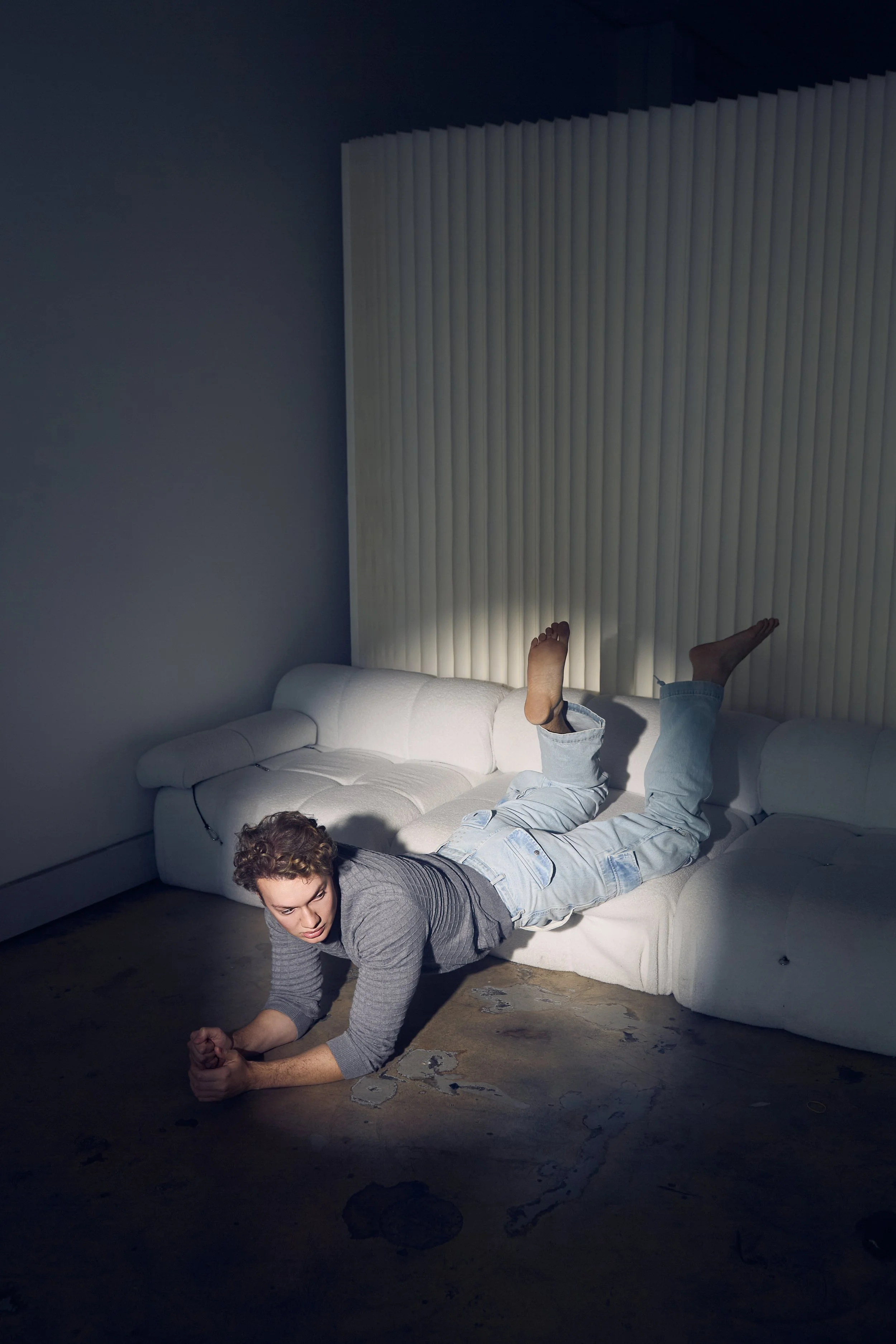 A man is lying on a white sofa with his head and upper body on the floor, and his legs raised onto the sofa. He is pausing on the floor in a plank position, facing downward with his palms and forearms on the floor. The room has a white wall with vert