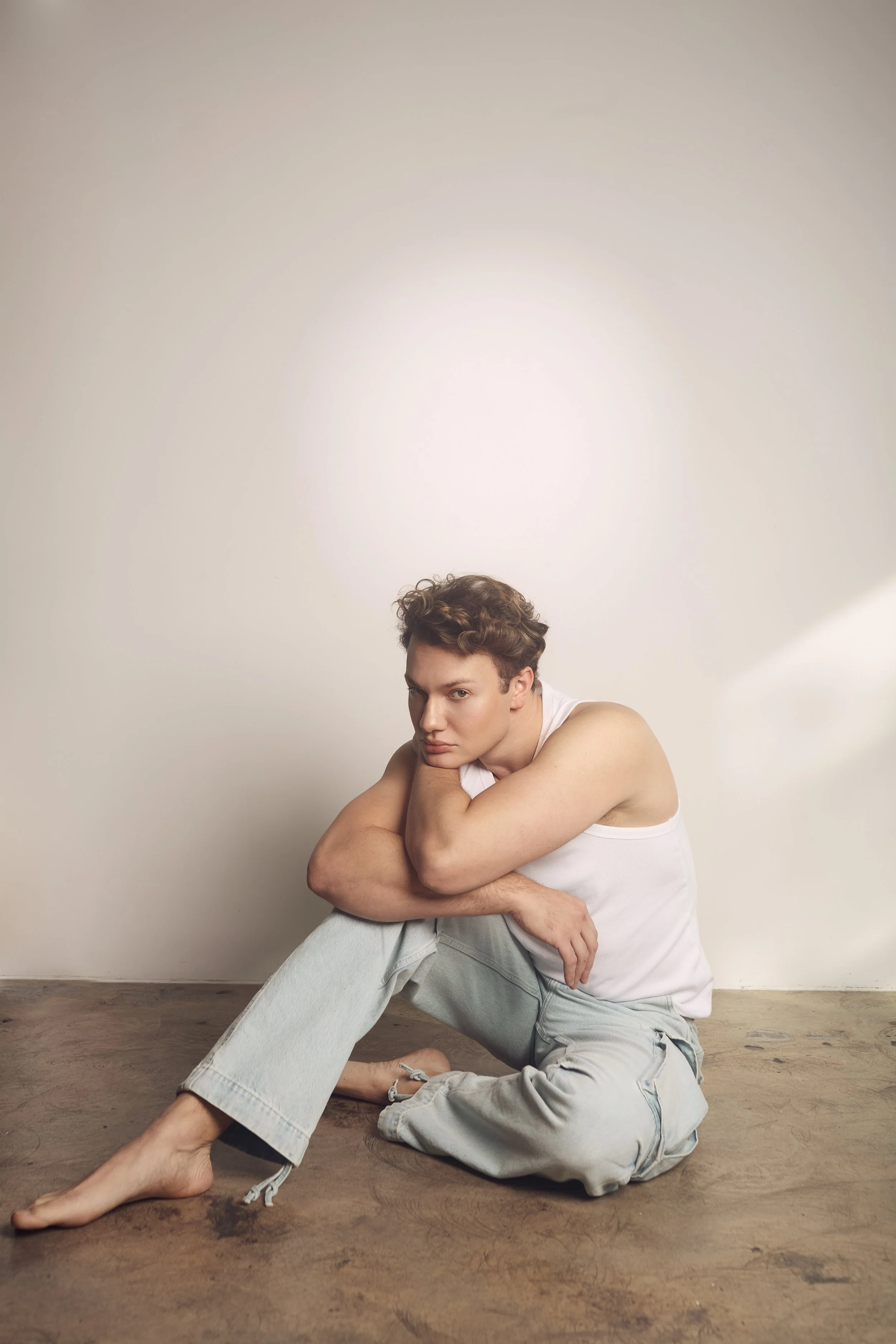 A young man with curly hair sits on the wooden floor with a neutral expression, wearing a white tank top and light gray jeans, leaning his head on his crossed arms in front of a plain off-white wall.