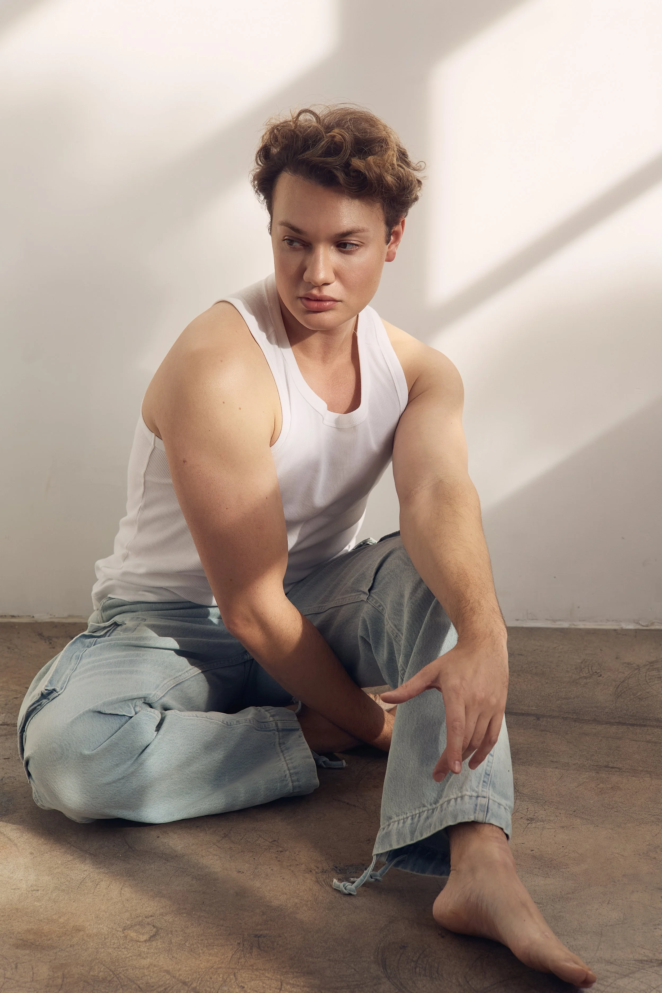 A young person with short, curly brown hair and light skin, wearing a white tank top and gray pants, sitting on the floor with arms crossed and looking down with a thoughtful expression against a plain white background with subtle shadows.