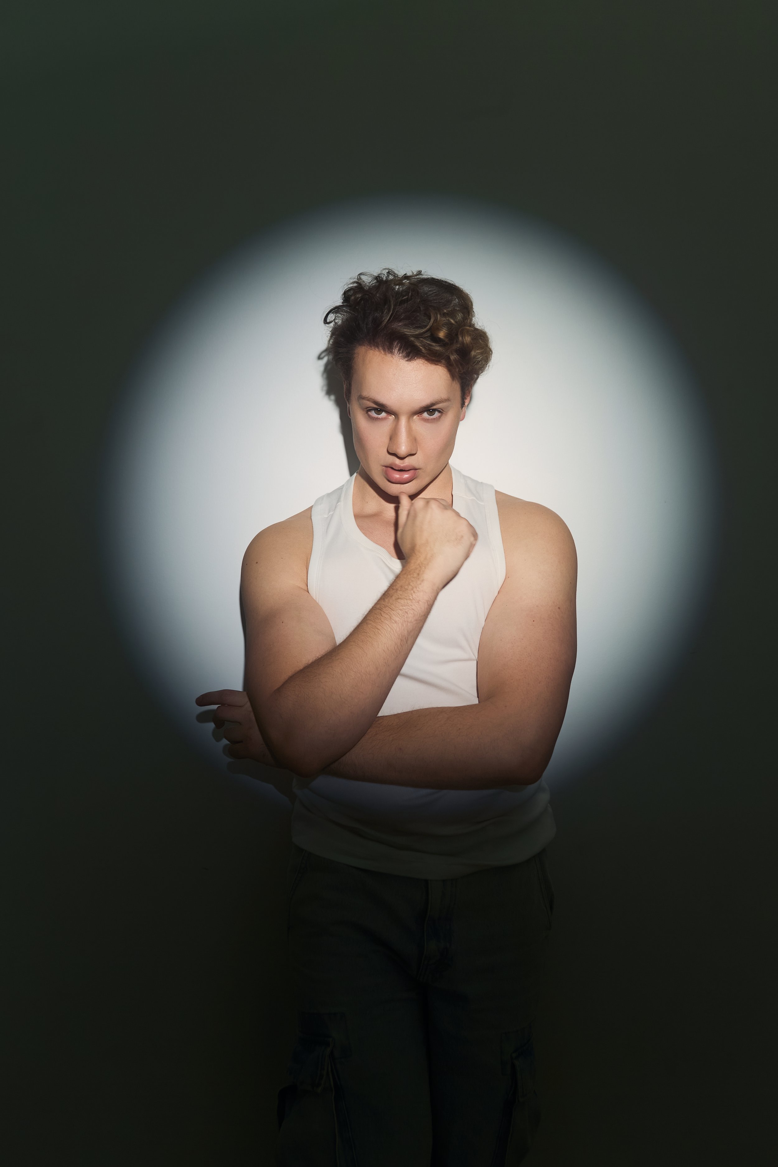 Young man with curly hair wearing a white sleeveless shirt standing in front of a spotlight with dark background, looking directly at the camera with a serious expression.