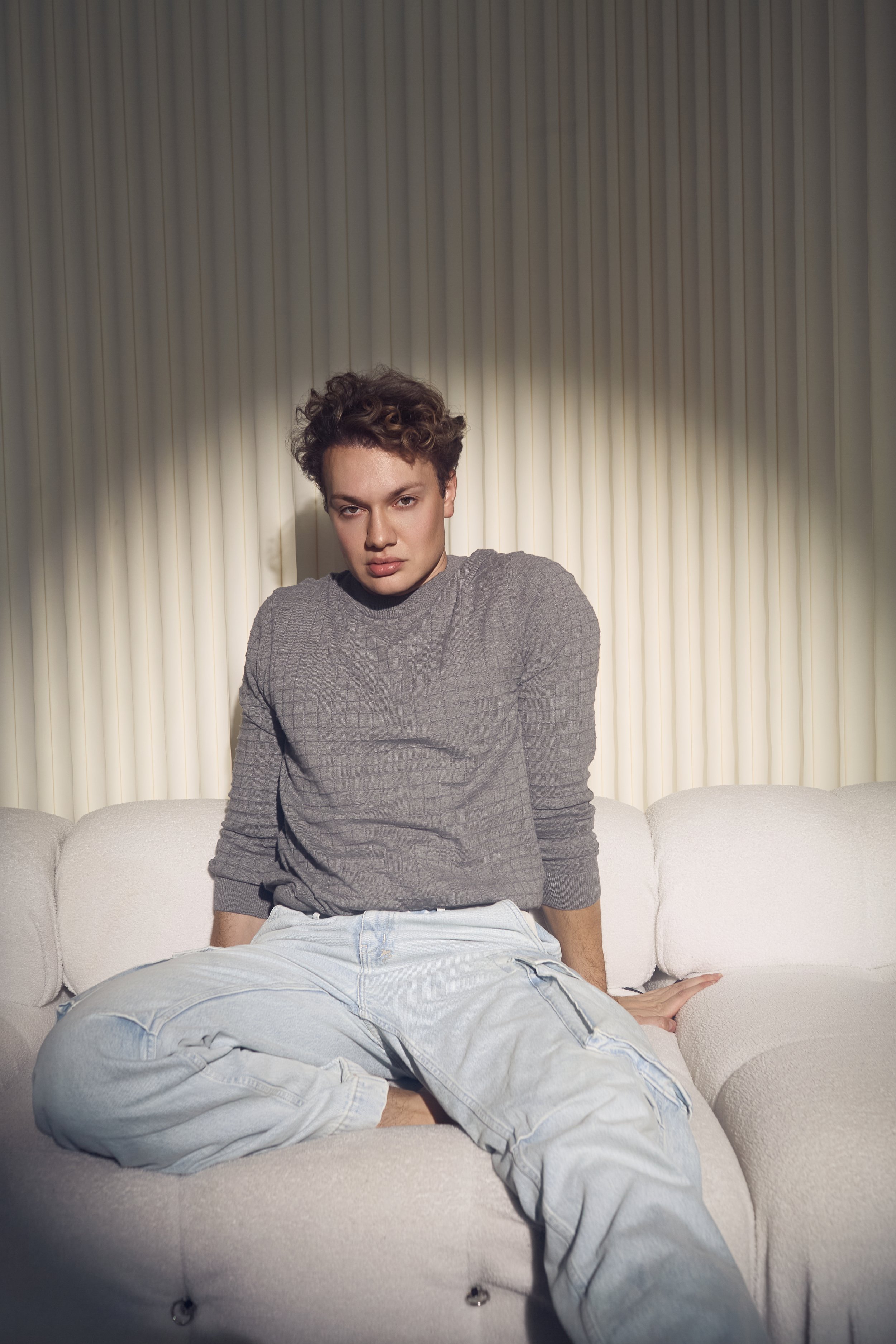 A woman with short, curly hair wearing a gray long-sleeve shirt and light-colored jeans, sitting on a beige sofa in front of a background with vertical blinds.