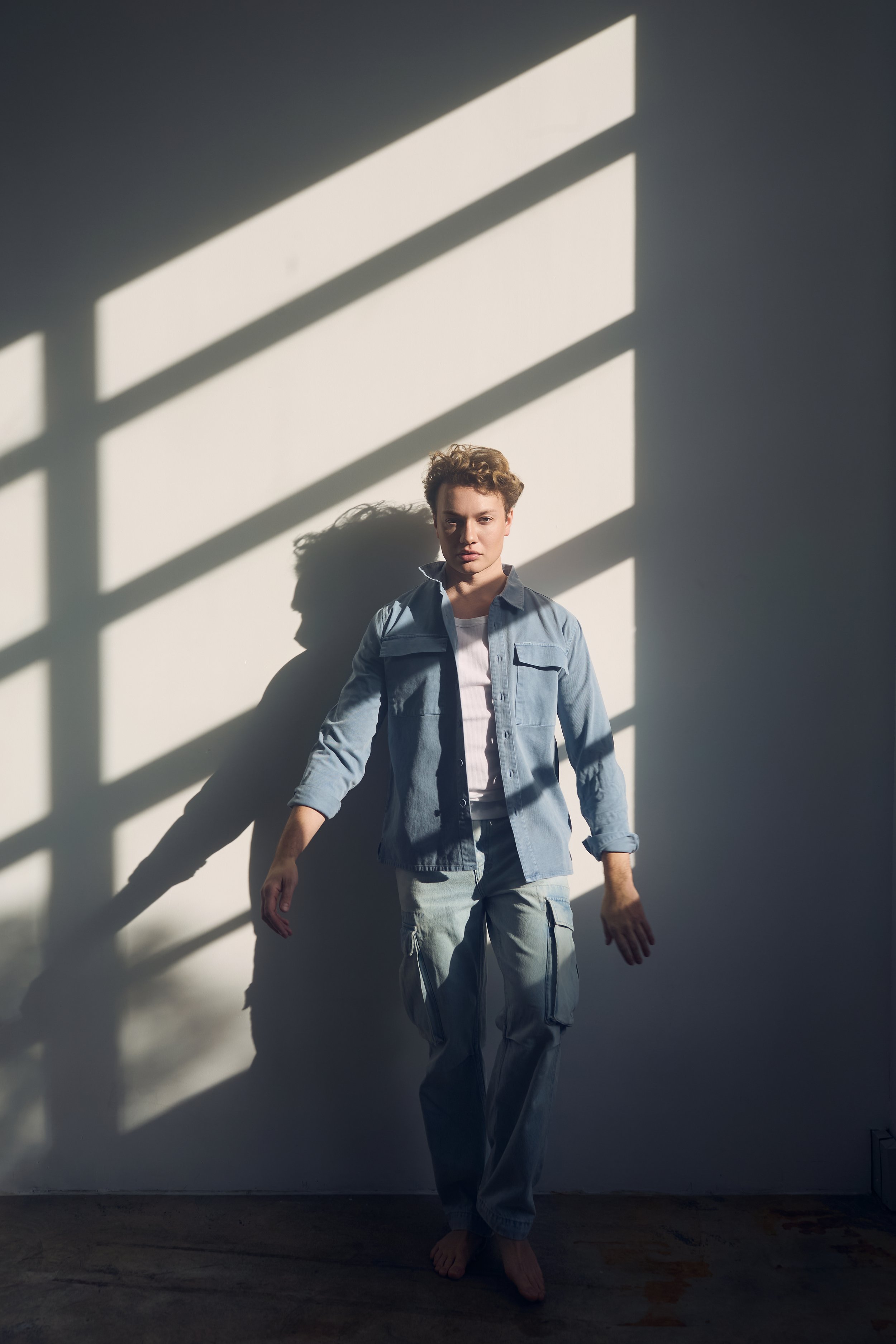 Young person with short curly hair standing barefoot against a white wall with shadows of a window cast across. Wearing light-colored pants and a denim shirt open over a white t-shirt.