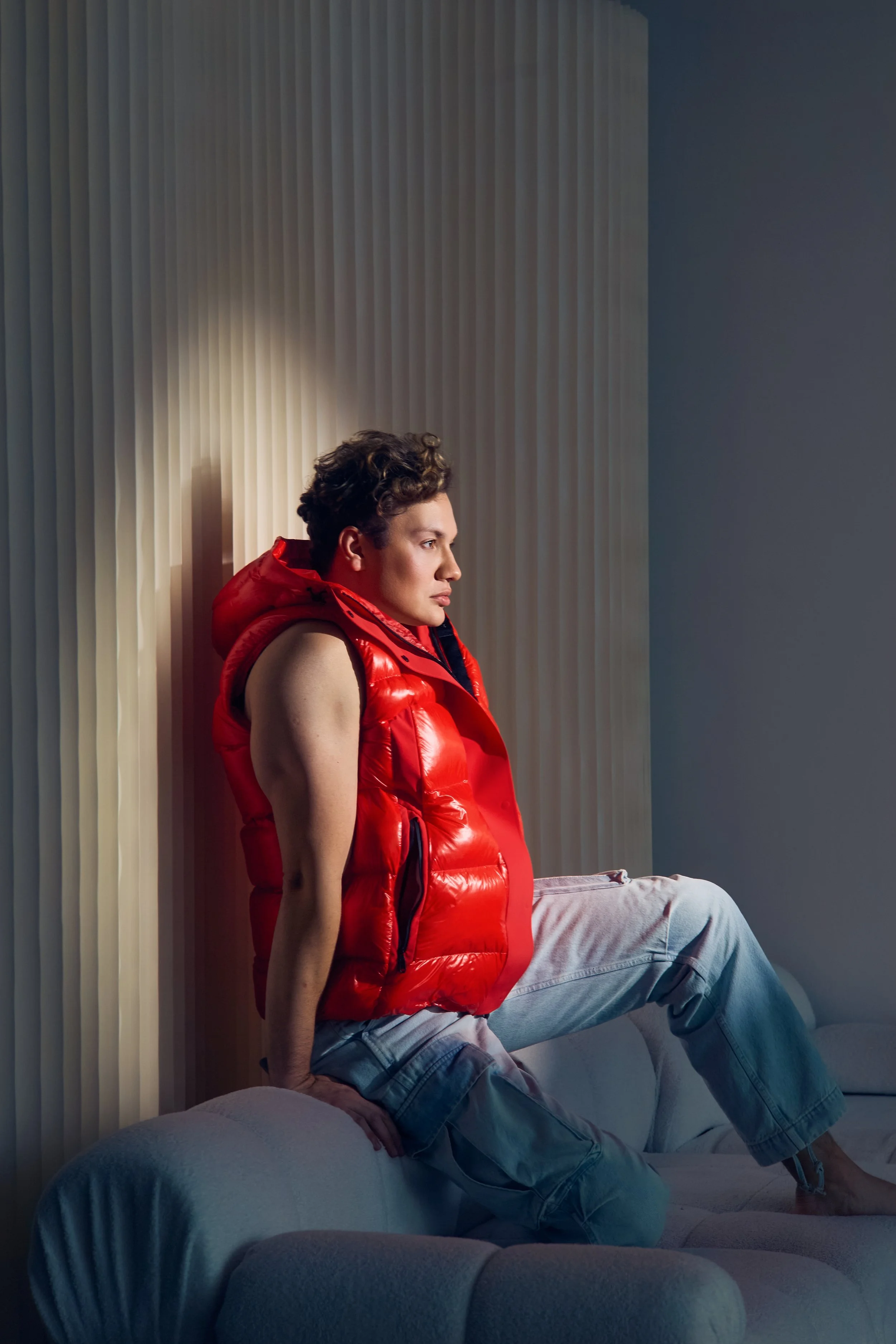 A young man with curly hair sitting on a sofa, wearing a red puffy vest and light-colored jeans, looking to the right with a serious expression, against a plain background with vertical blinds.