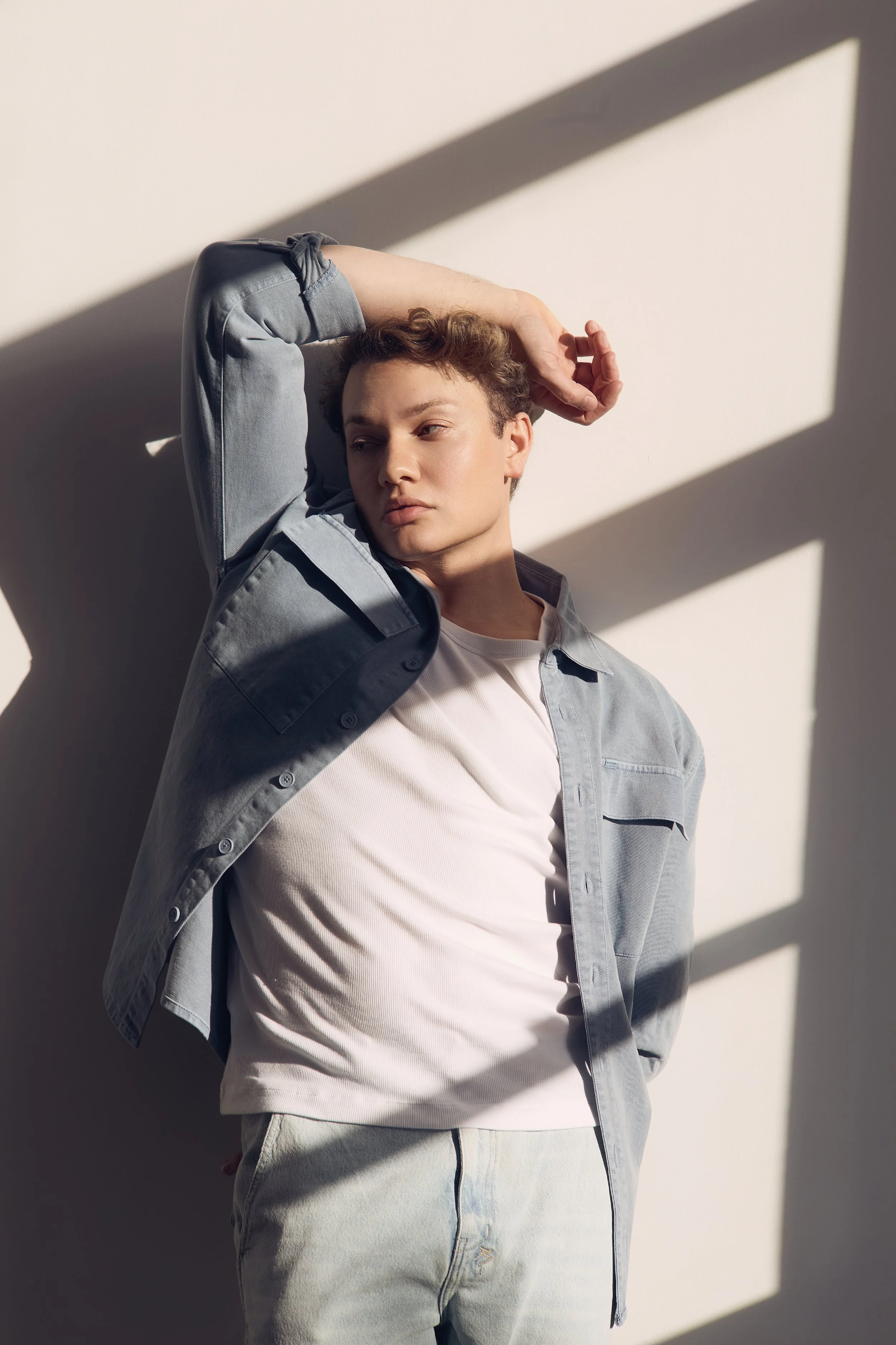Young man in casual gray denim shirt and light jeans leaning against a white wall, with sunlight casting shadows through window panes.