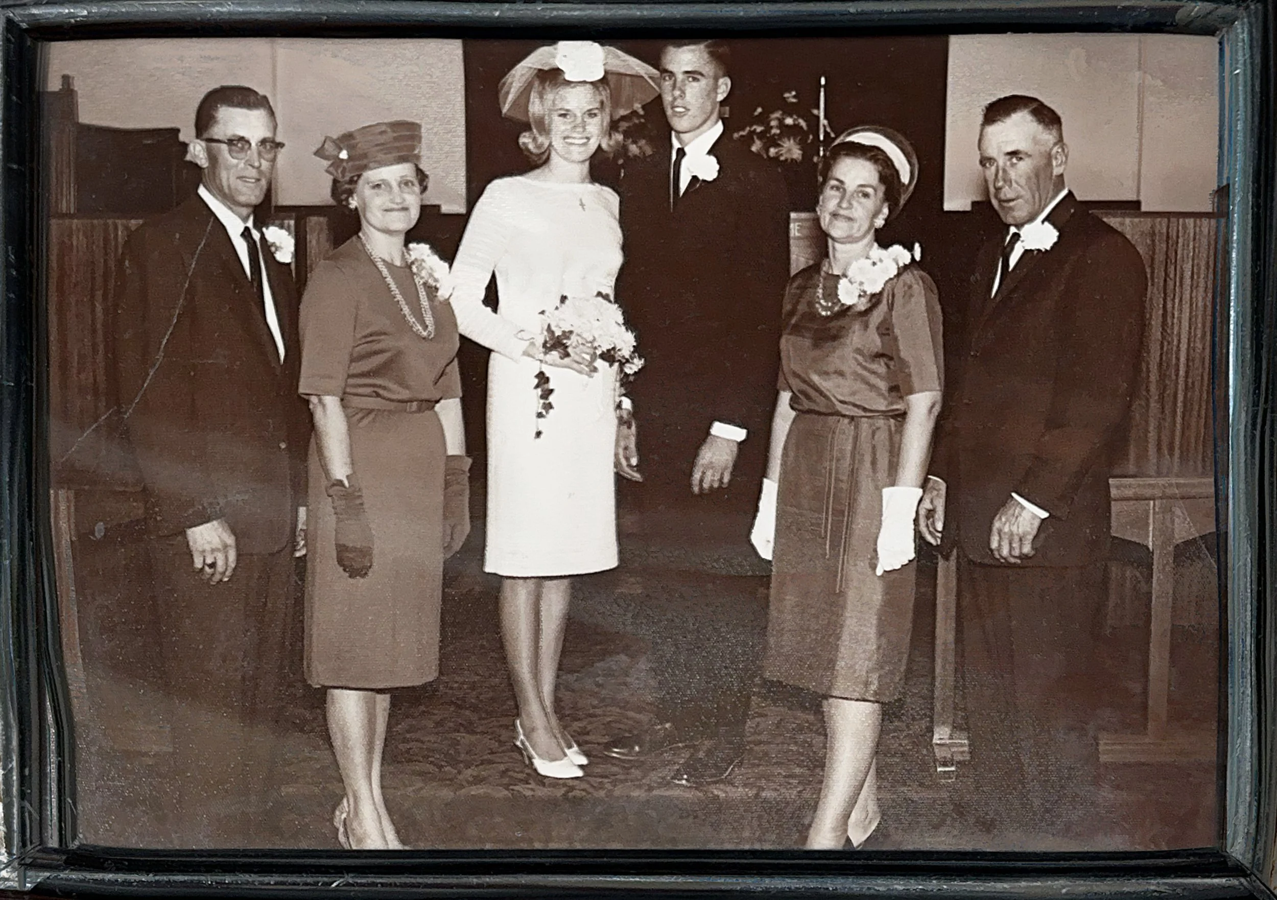 A black-and-white vintage wedding photo showing a couple with four other people, all dressed in formal attire. The bride is wearing a white dress and holding a bouquet, with a smile on her face. The groom and other people are dressed in dark suits or dresses, with corsages and corsages. The background suggests an indoor setting, possibly a church or reception hall.