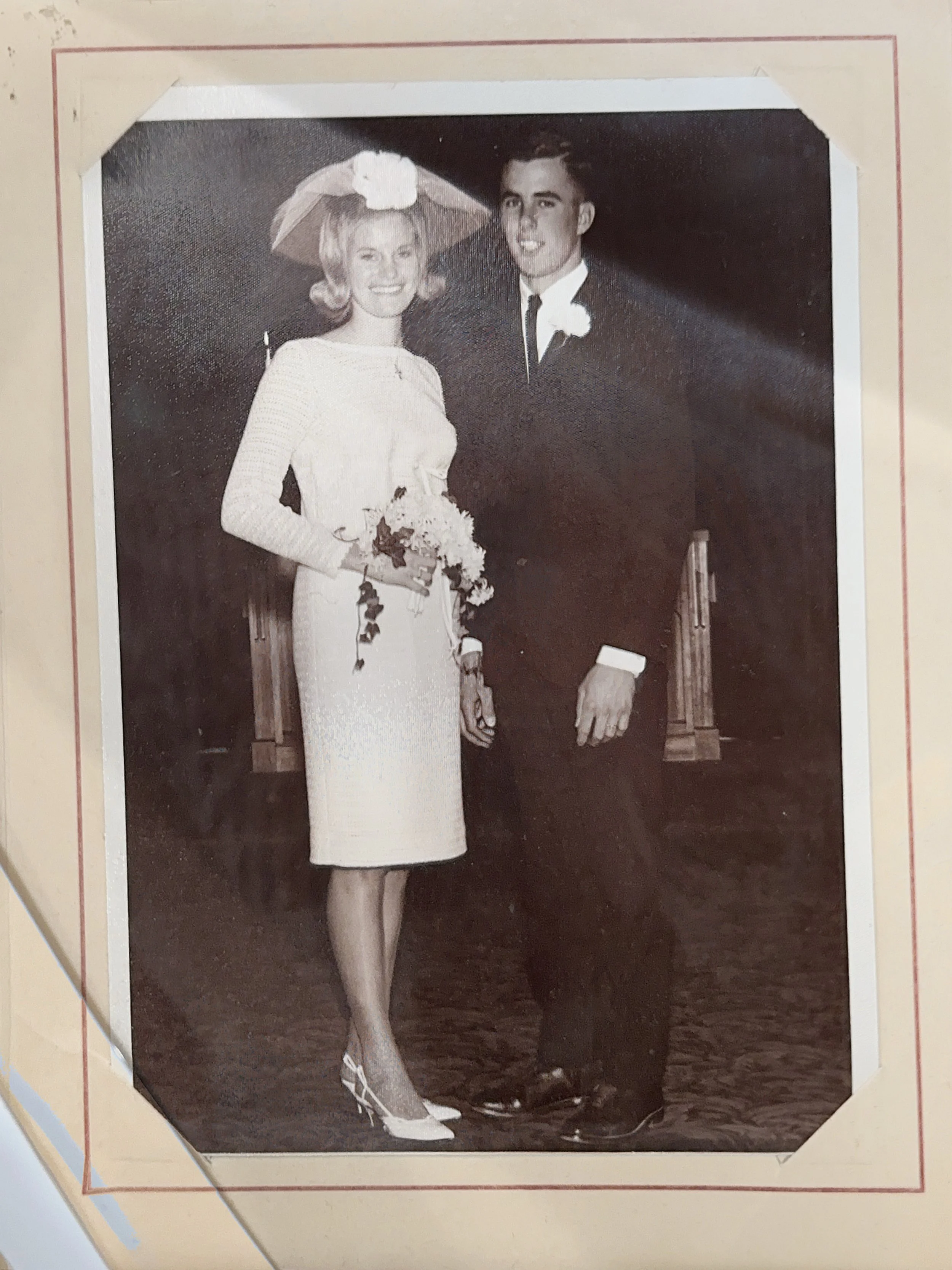Black and white photograph of a young woman and a young man dressed in formal attire; the woman is wearing a white dress and hat with a veil, holding flowers, and the man is in a tuxedo with a boutonnière.