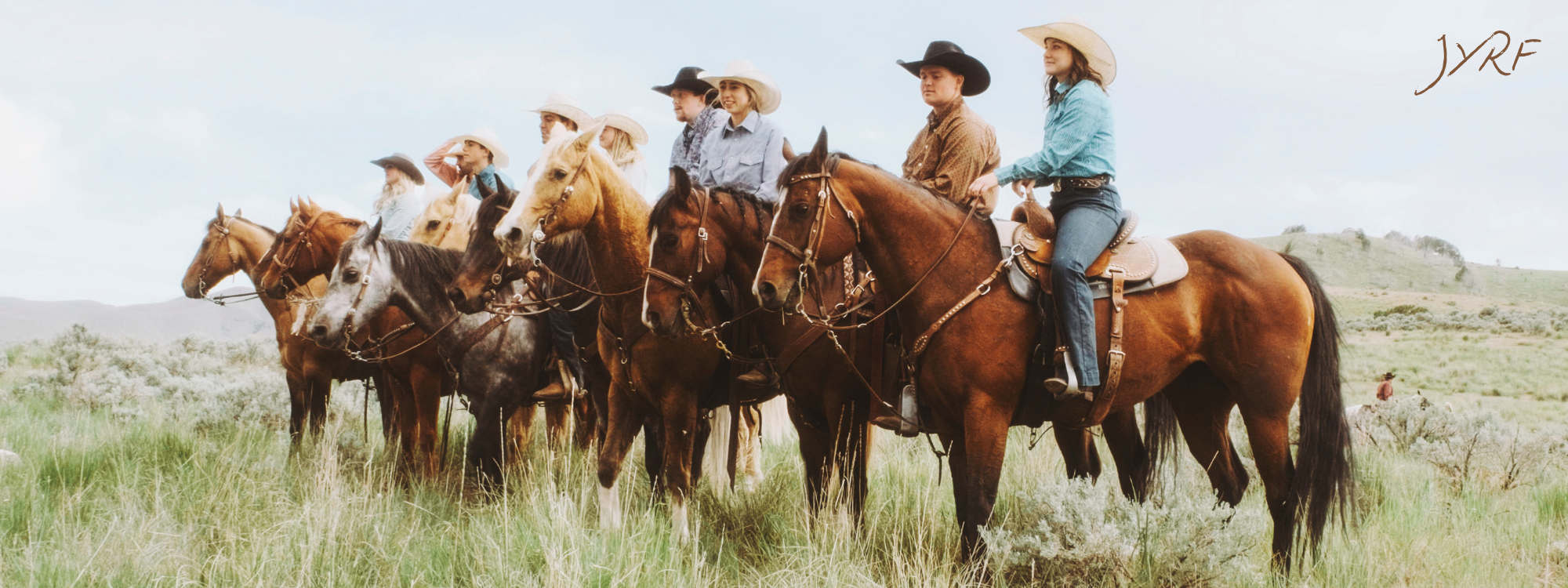 Group of people riding horses in an open grassy field, wearing western hats and casual Western attire, with a hilly landscape in the background.