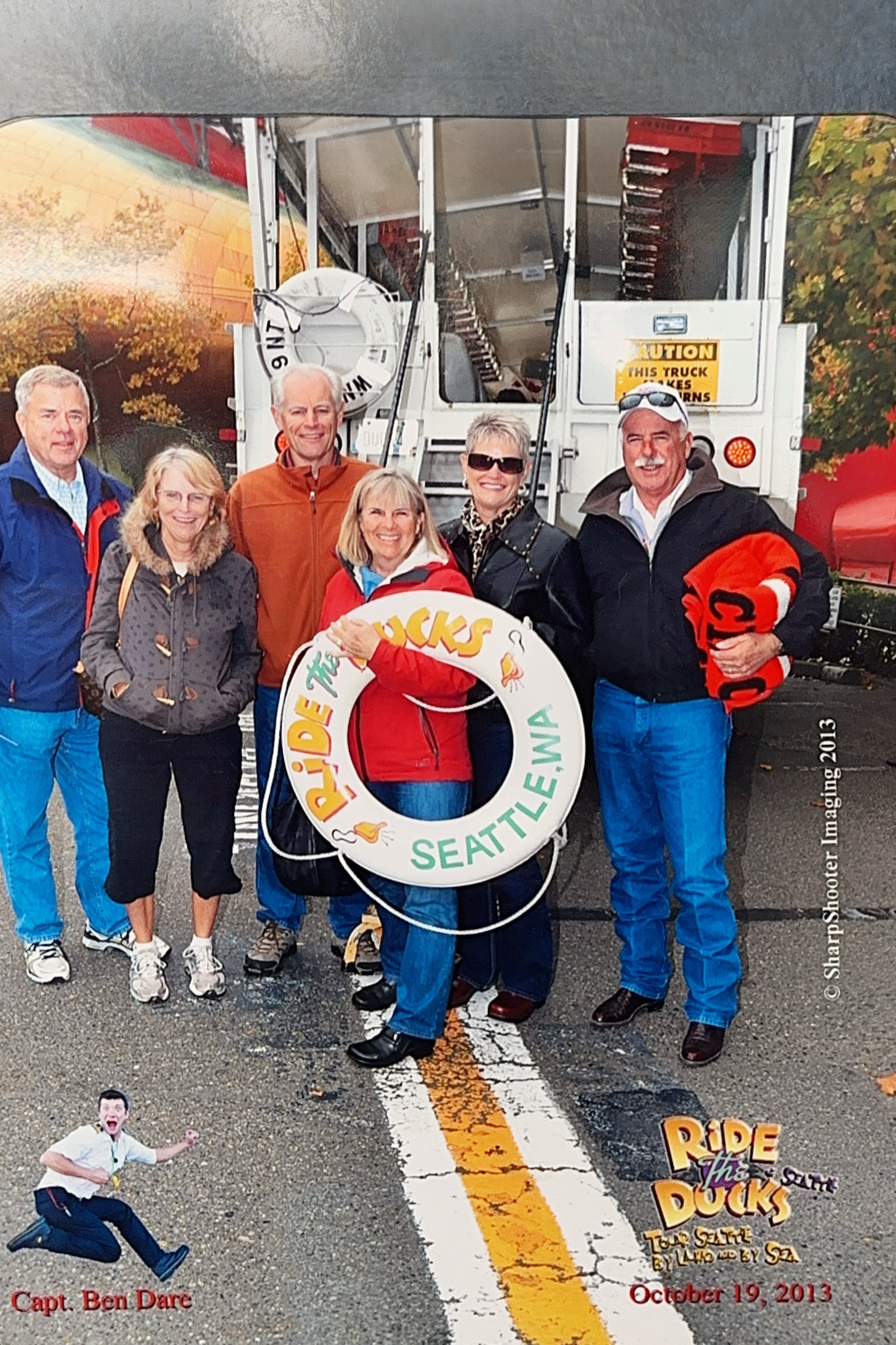 Group of people posing outdoors, some holding a Life Ring, with a truck and autumn trees in the background. The image is part of promotional material for an event called Ride the Ducks in Seattle, dated October 19, 2013.