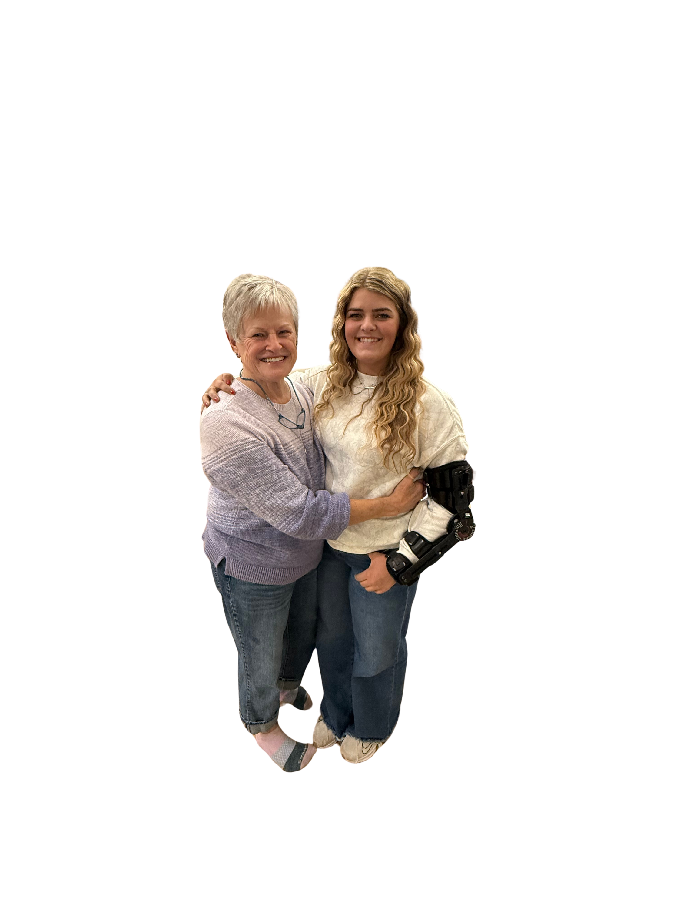 A woman with short gray hair and a young woman with long wavy blonde hair smiling and hugging each other, standing close together, dressed casually, against a black background.