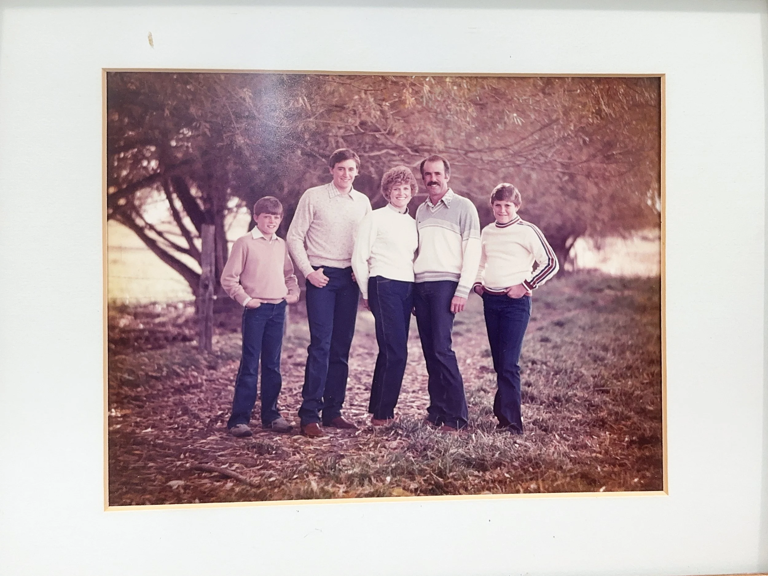 Family of five standing outdoors on a path covered with fallen leaves, with trees in the background and warm autumn lighting.
