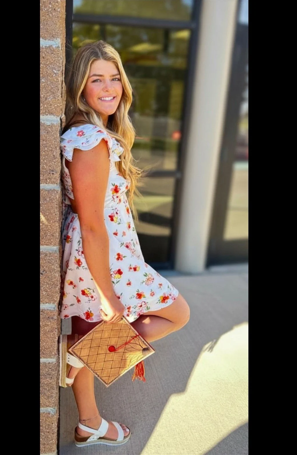 A young woman with long blonde hair wearing a white dress with red and yellow floral print, leaning against a brick wall outside a building. She is smiling, holding a small brown graduation cap with a red tassel in her right hand, and wearing white sandals.