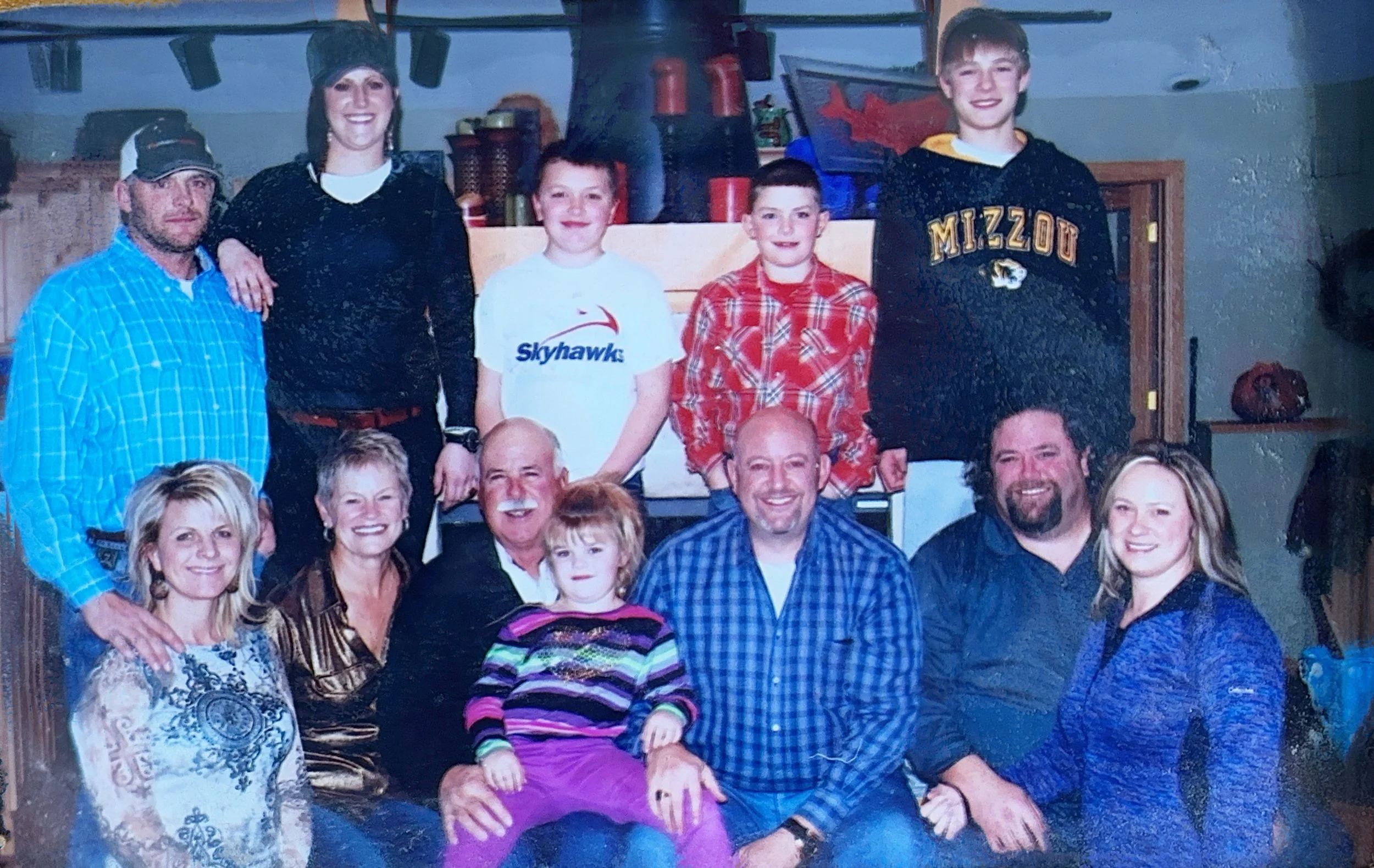 A large group of people including children, teenagers, and adults posing indoors, with some standing and some sitting. The background shows shelves with cups and containers. Most individuals are smiling.