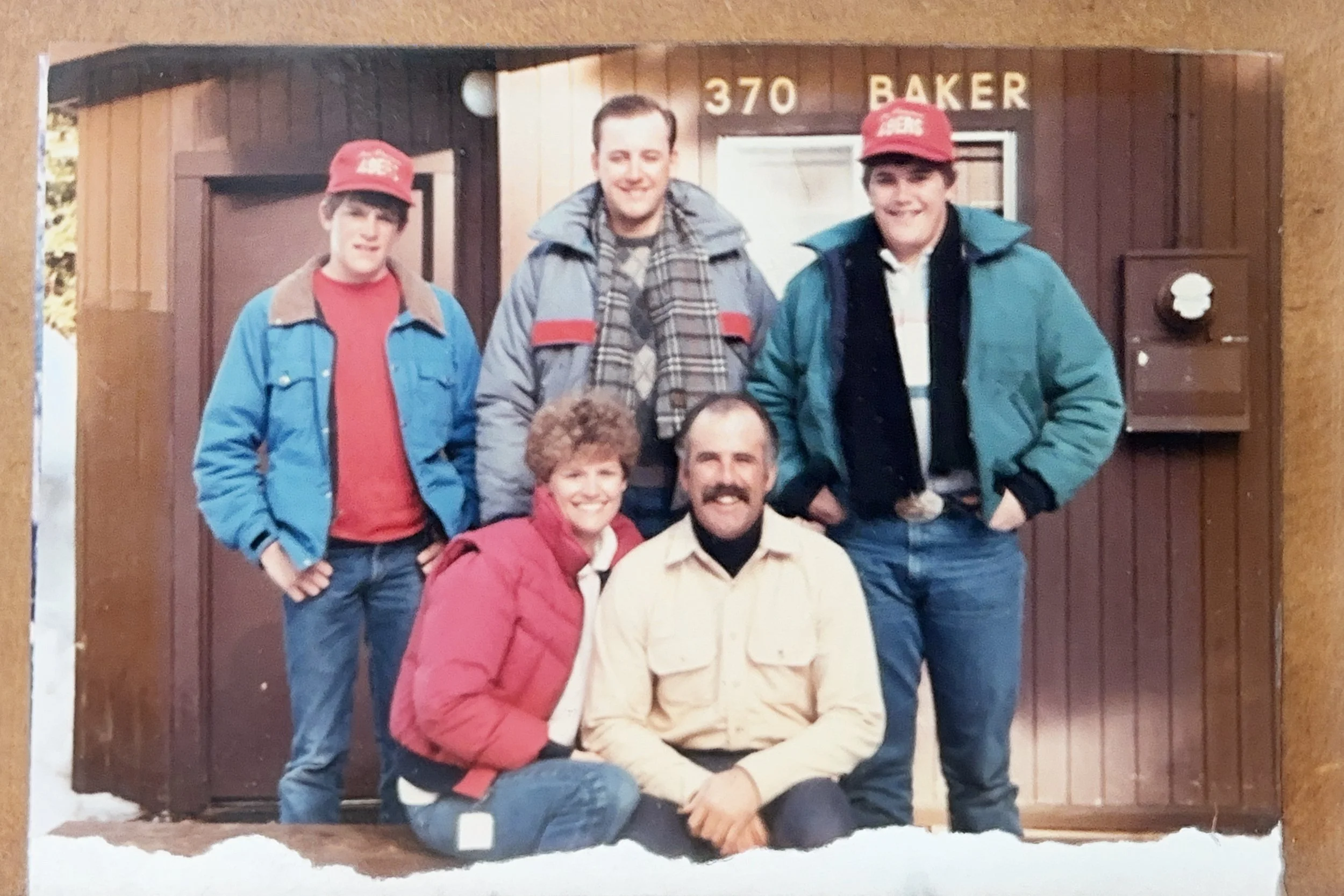 A group of five people, three standing and two sitting, posing outdoors in front of a wooden building labeled '370 BAKER'. The group consists of two young men in red hats, an older woman in a red jacket, an older man in a beige jacket, and a man standing between the two young men in a gray jacket. All are smiling.