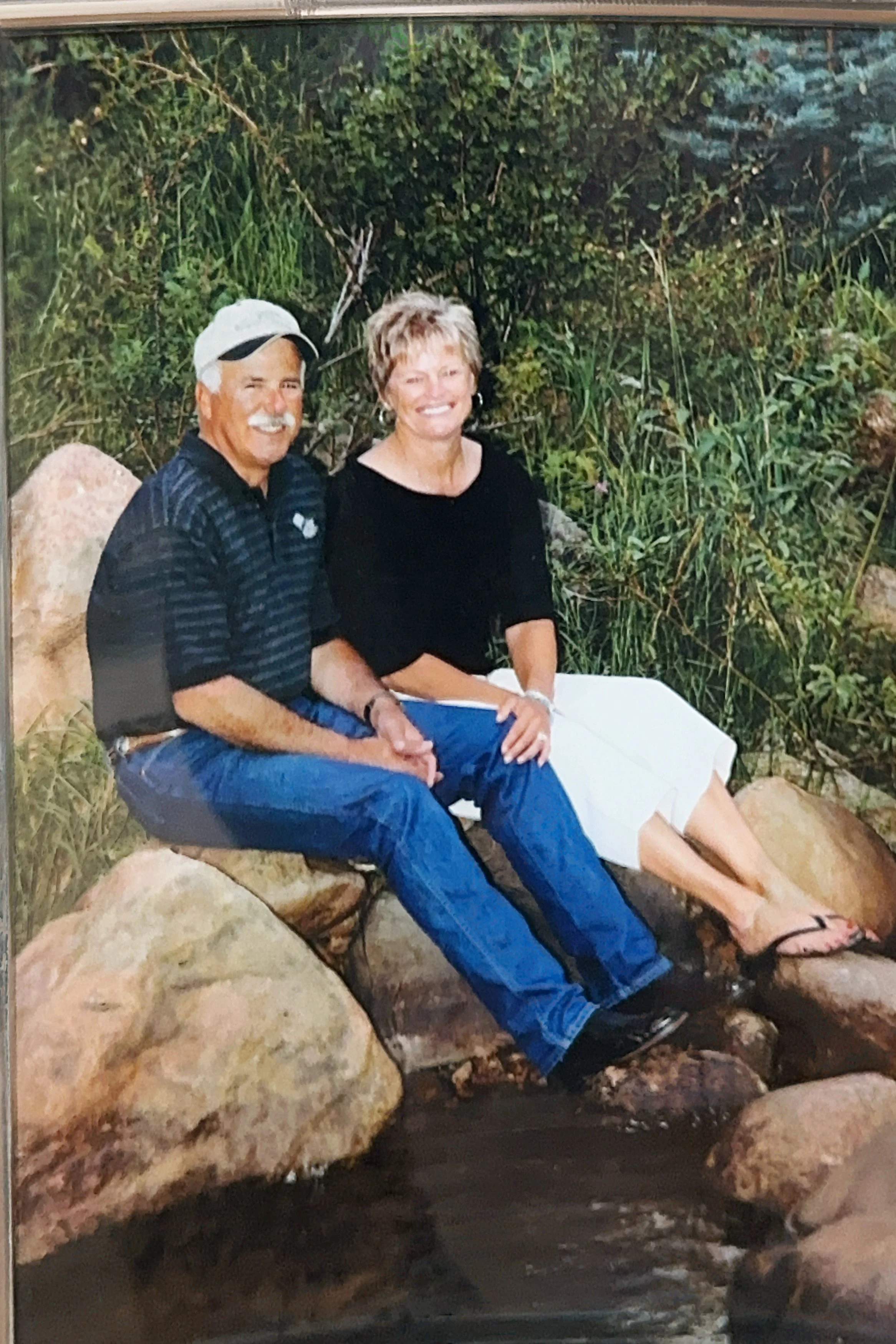 A man and woman sitting on rocks by a small waterfall surrounded by green plants and trees, smiling at the camera.