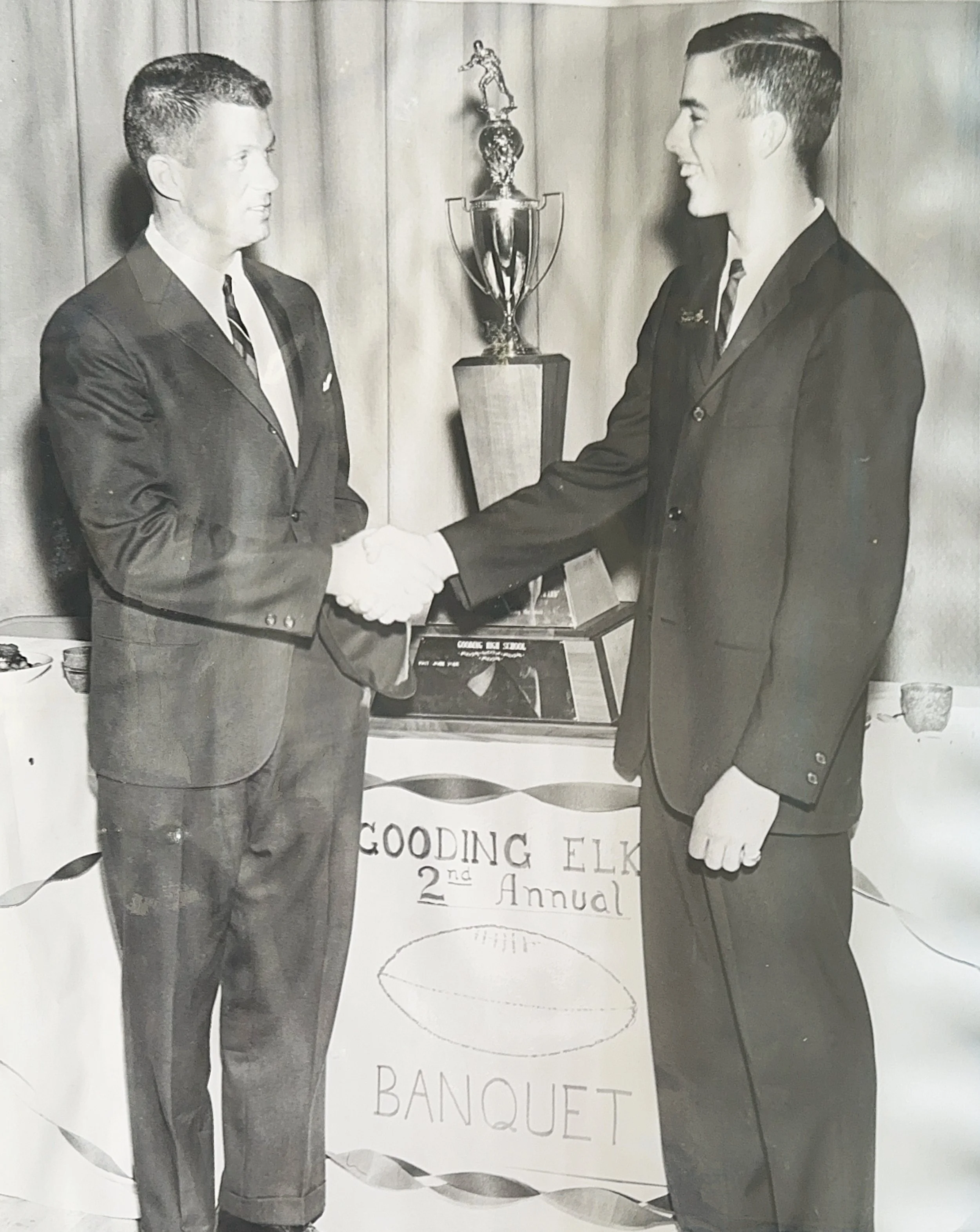 Black and white photo of two men in suits shaking hands at a banquet, with a large trophy and a sign reading 'Gooding Elk 2nd Annual' in the background.