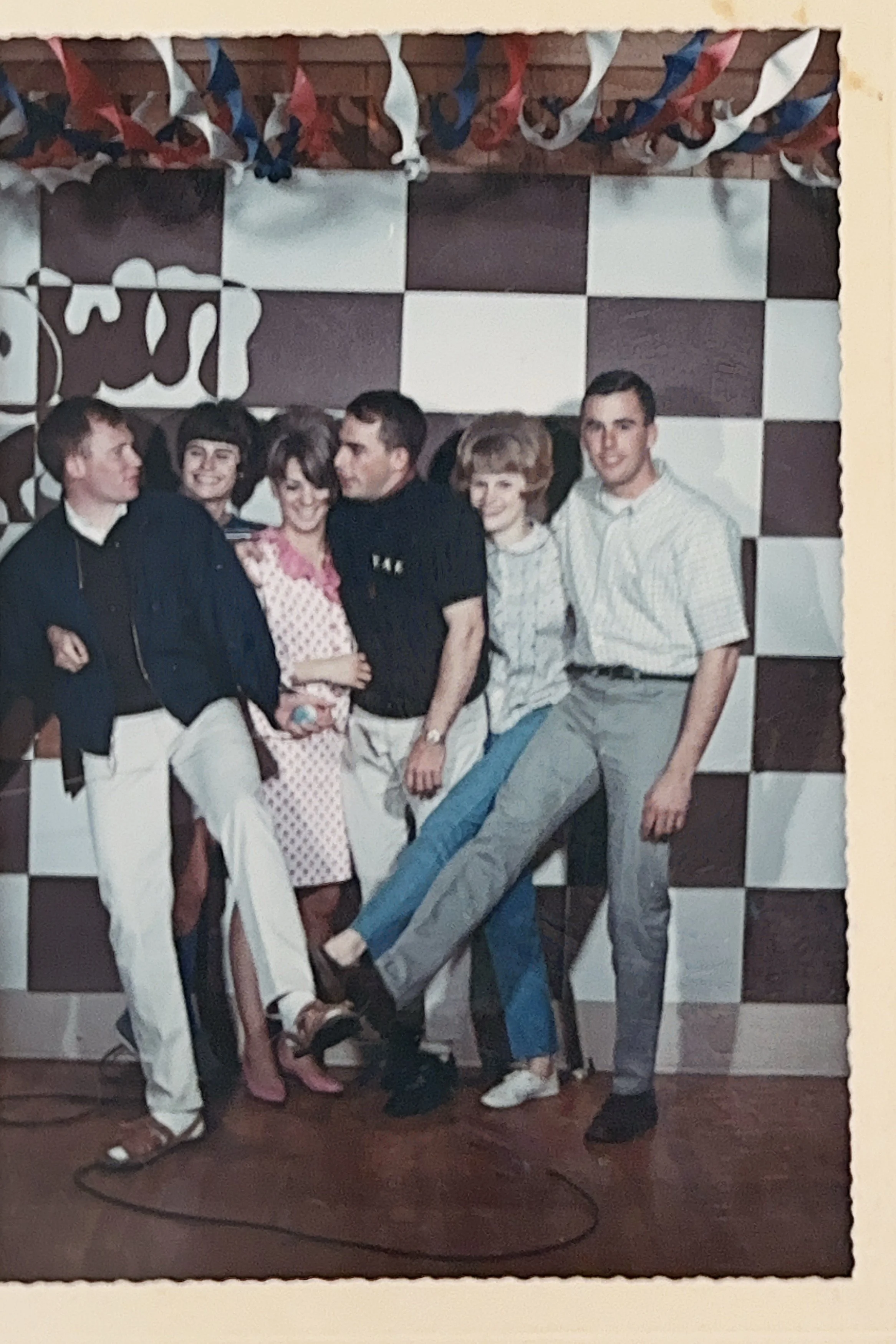 A group of six young adults posing together in front of a black-and-white checkered wall with party streamers hanging from the ceiling.